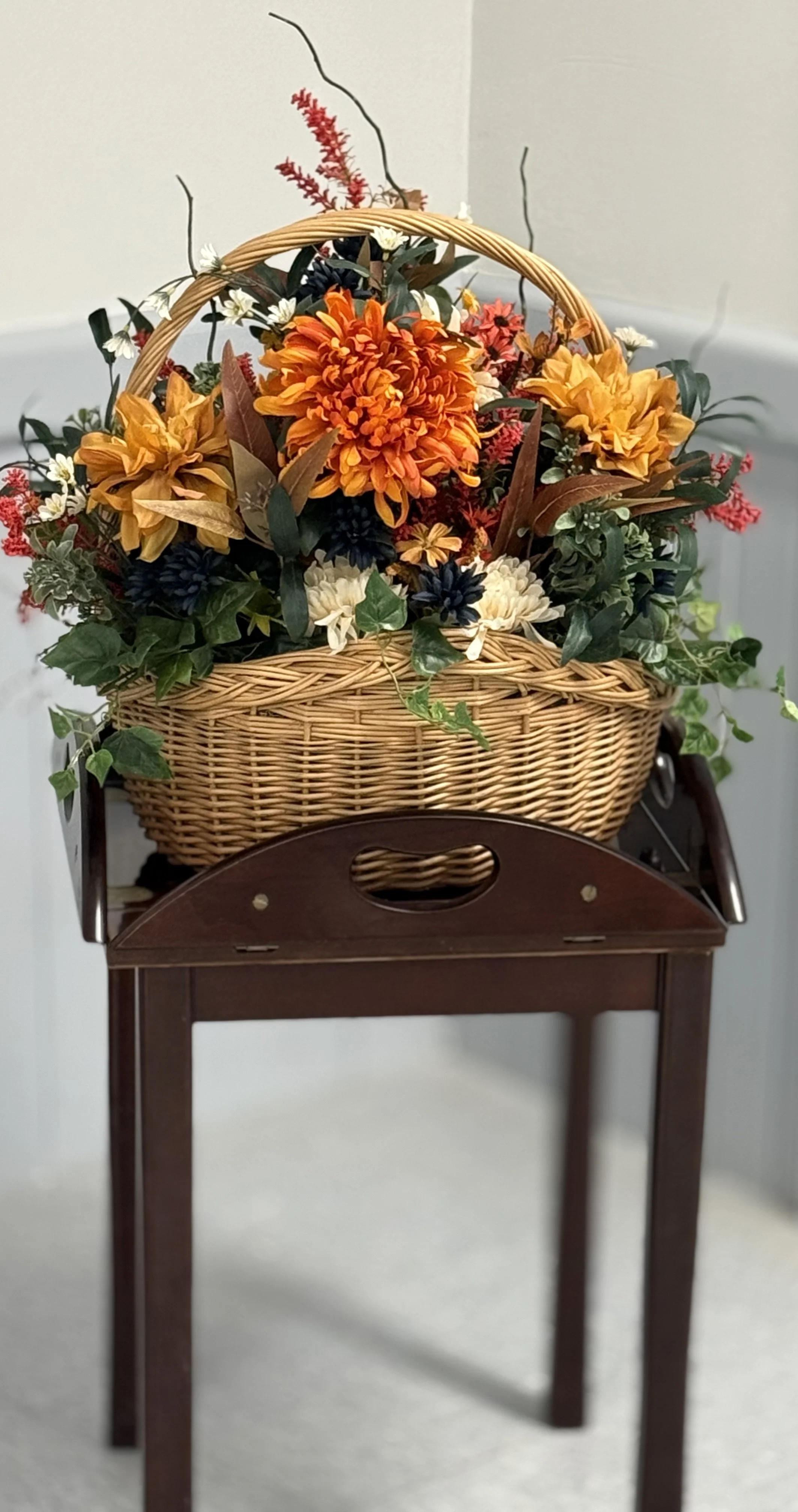 A wicker basket filled with orange, white, and dark purple artificial flowers, with green leaves, placed on a wooden stand.