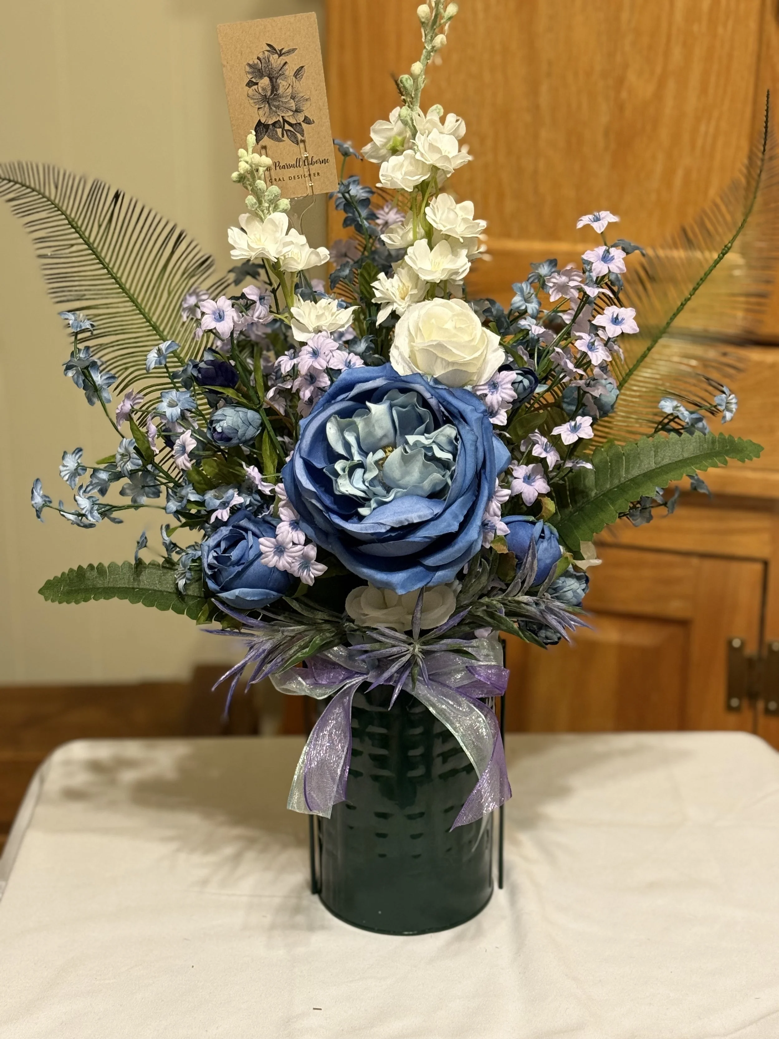 A floral arrangement with white, purple, and blue flowers in a black vase, decorated with a purple ribbon, placed on a table.