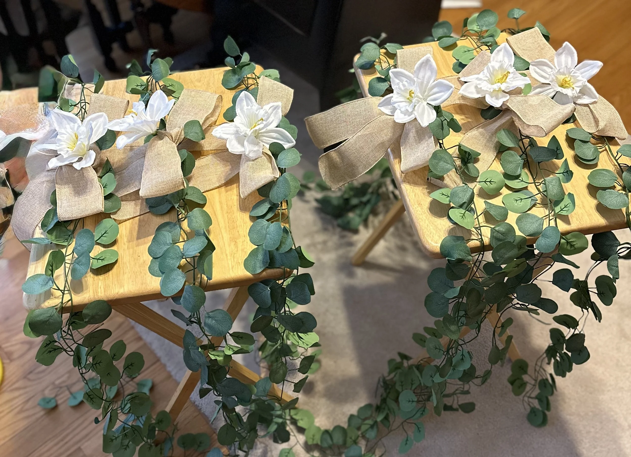 Two wooden chairs decorated with beige bows, white flowers, and green eucalyptus garlands.