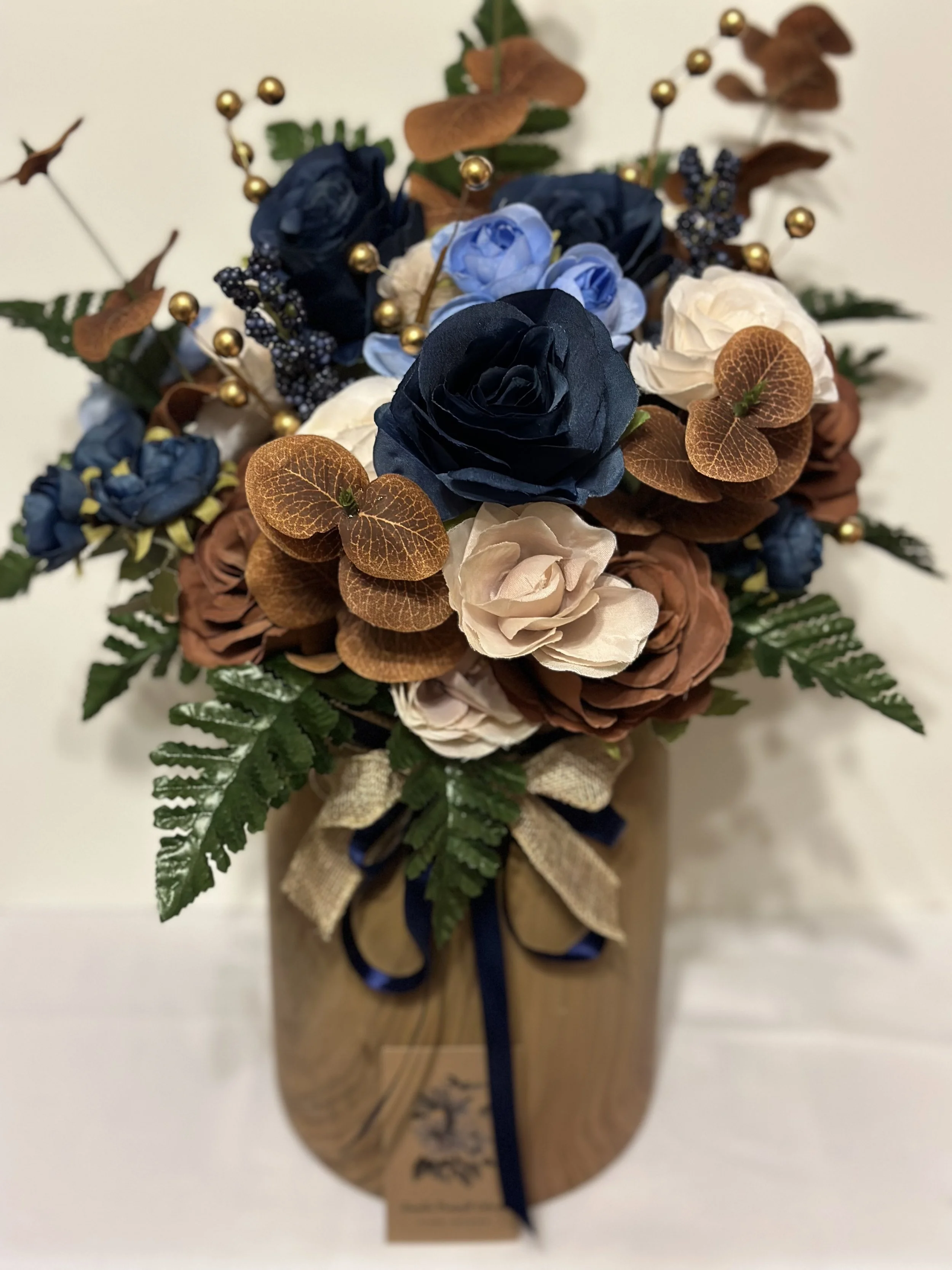 A bouquet of artificial flowers with blue, white, and brown roses, decorated with golden beads and green ferns in a wooden vase.