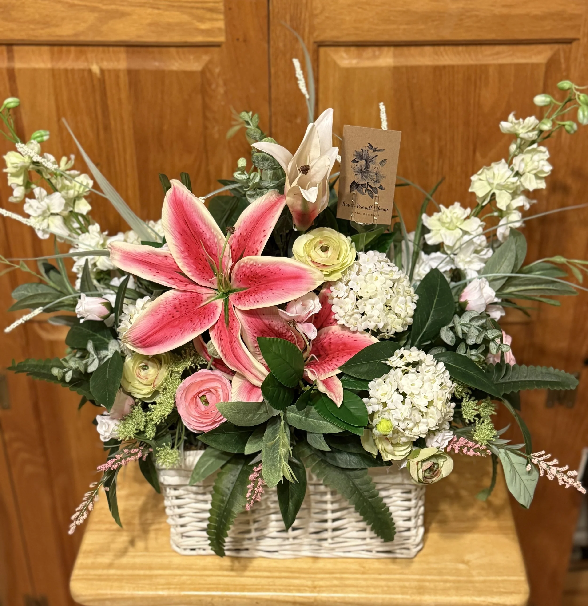 A floral arrangement in a white wicker basket featuring pink and white lilies, roses, hydrangeas, and assorted greenery, set against a wooden background.