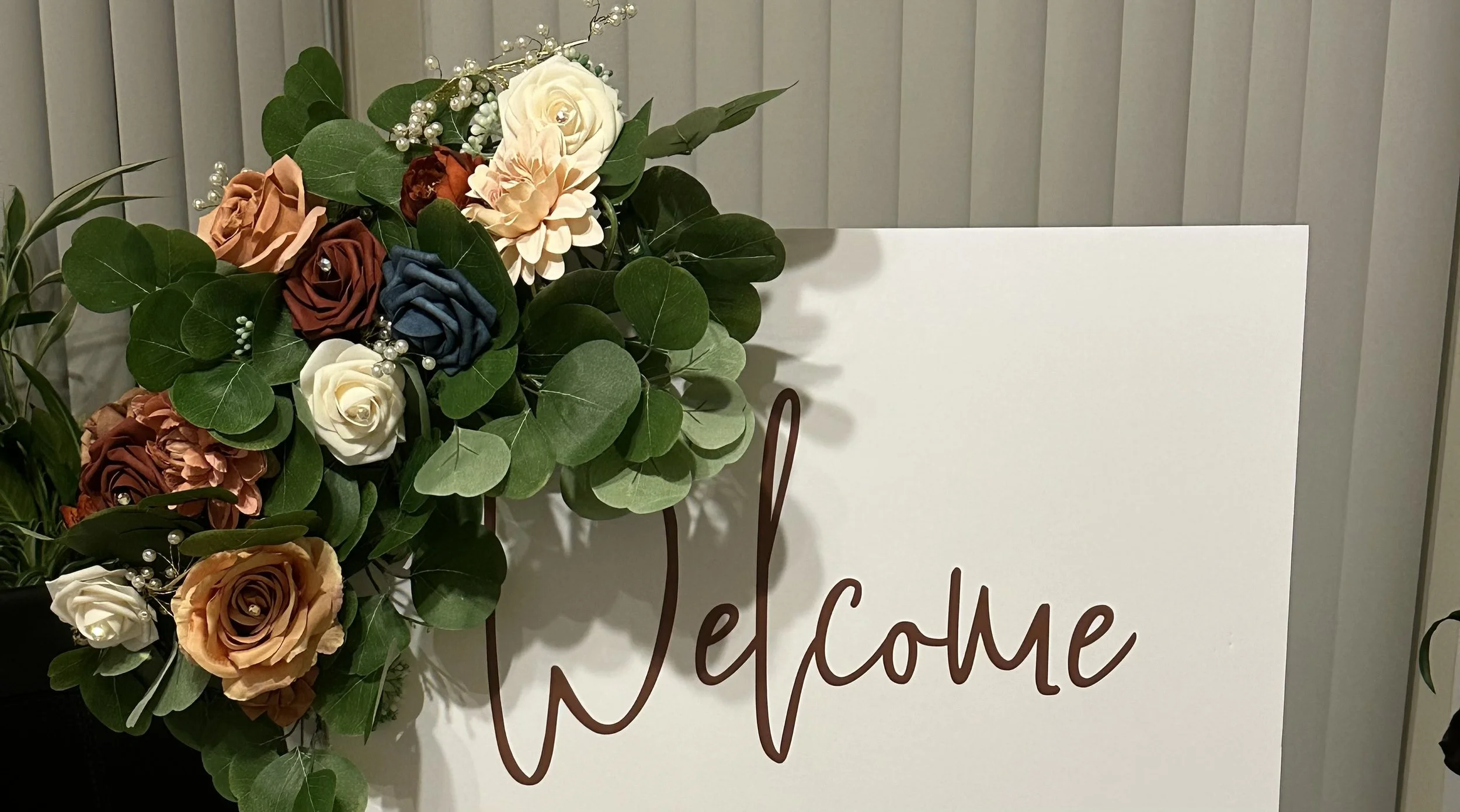 A floral arrangement of roses, eucalyptus, and decorative flowers next to a white welcome sign with brown cursive lettering.