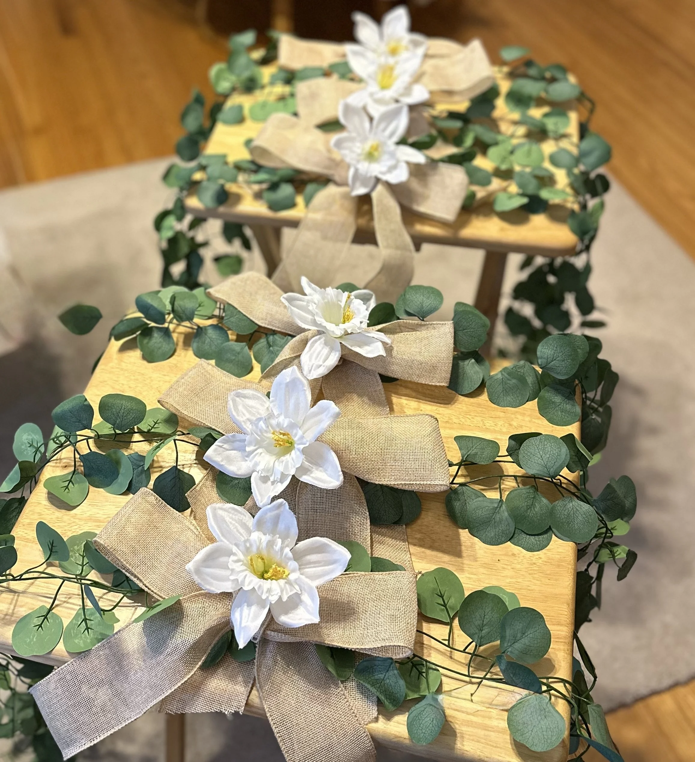 Decorative arrangement with white flowers, beige bows, and green eucalyptus leaves on wooden tables.