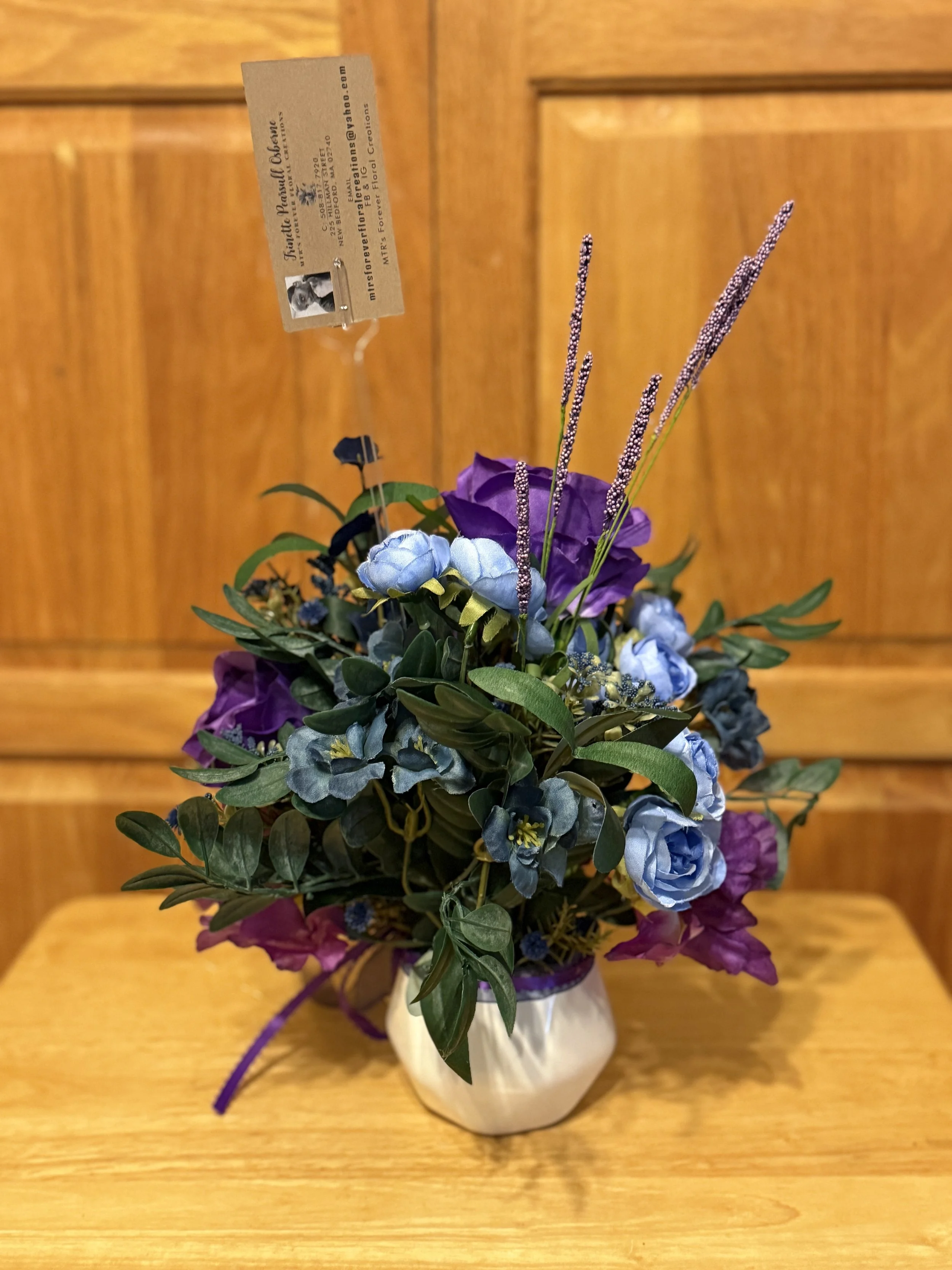 A floral arrangement with purple and blue flowers in a white vase on a wooden table against a wooden background.
