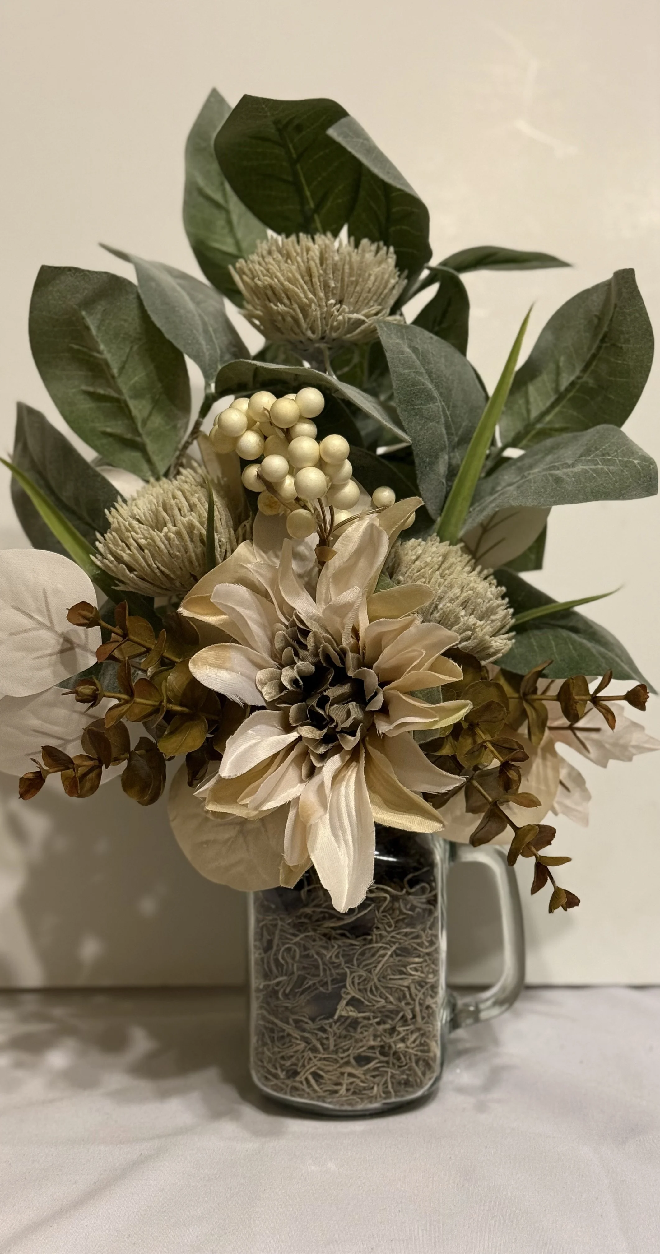 A bouquet of artificial flowers in a glass mug-shaped vase, including large cream-colored flowers, green leaves, white berries, and dried filler flowers, arranged on a white surface against a plain background.