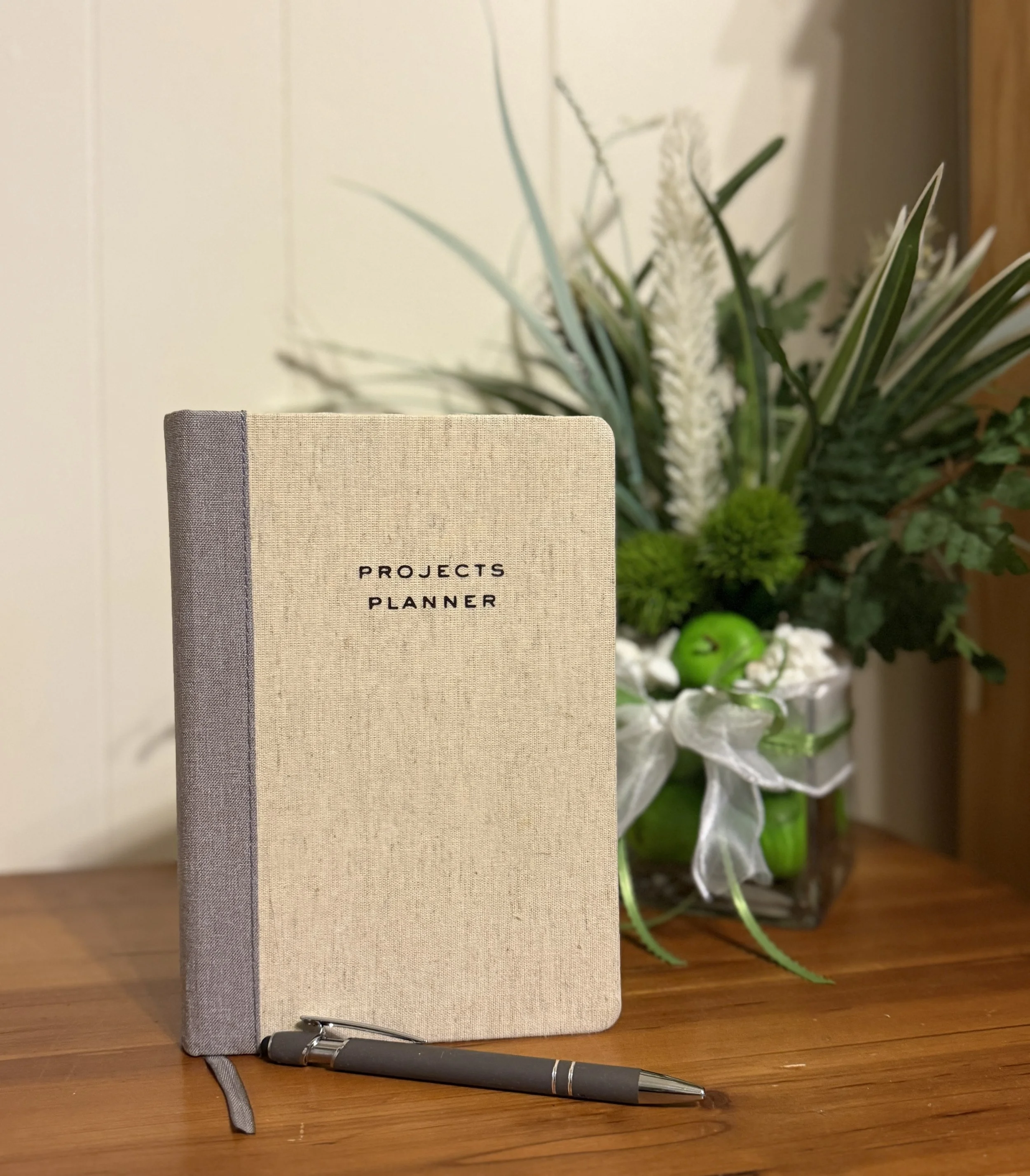 A beige hardcover planner with the words 'PROJECTS PLANNER' printed in black, placed on a wooden surface beside a silver pen with black grip. In the background, a glass vase containing green apples and green plants and white wheat flower centered.