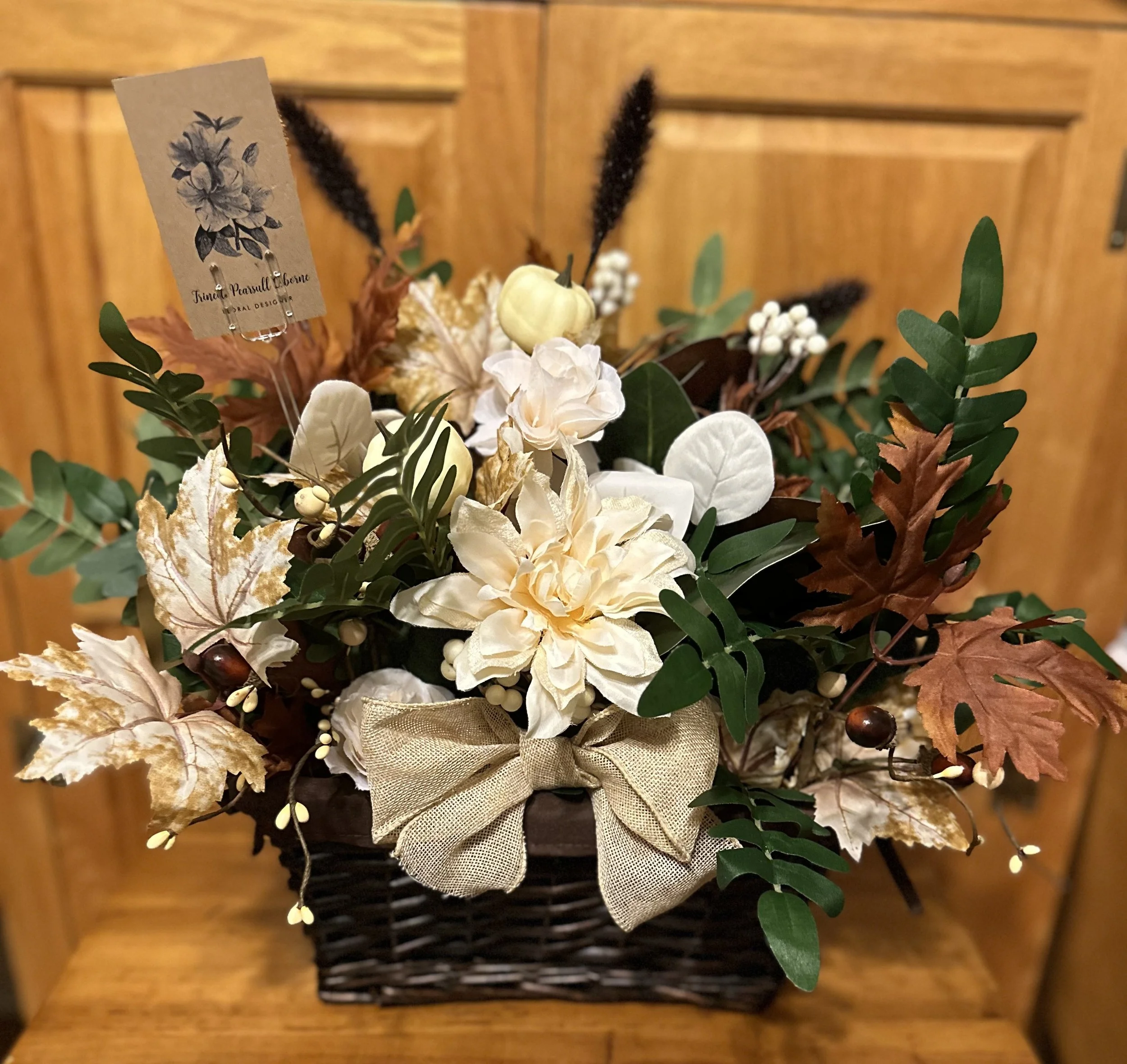 A decorative flower arrangement in a woven basket with white and cream flowers, green leaves, dried brown leaves, and a beige ribbon bow, placed on a wooden surface with a wooden background.