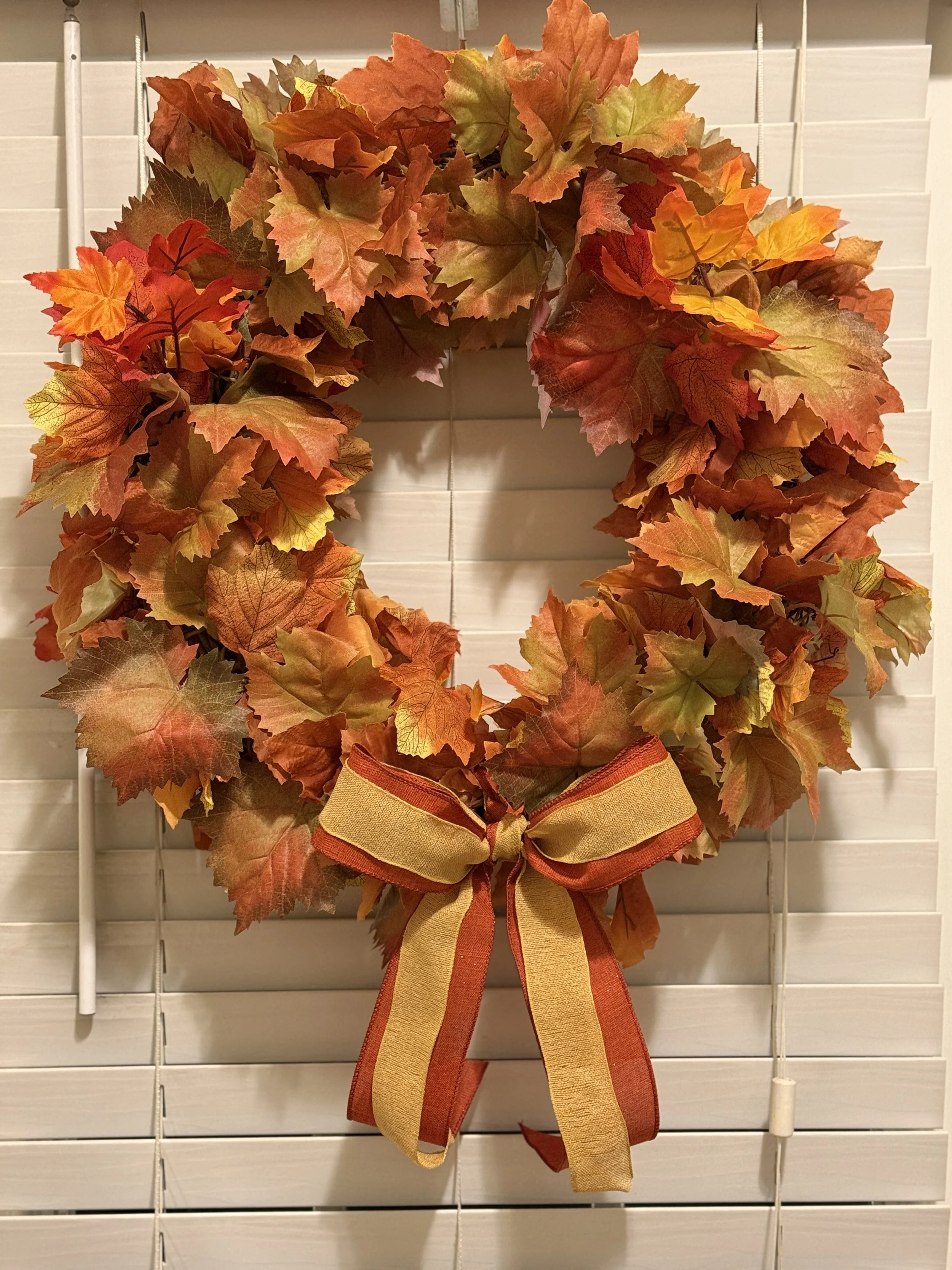 Autumn-themed decorative wreath made of orange, red, and yellow artificial leaves, adorned with a matching orange and yellow striped bow, hanging against white wooden blinds.