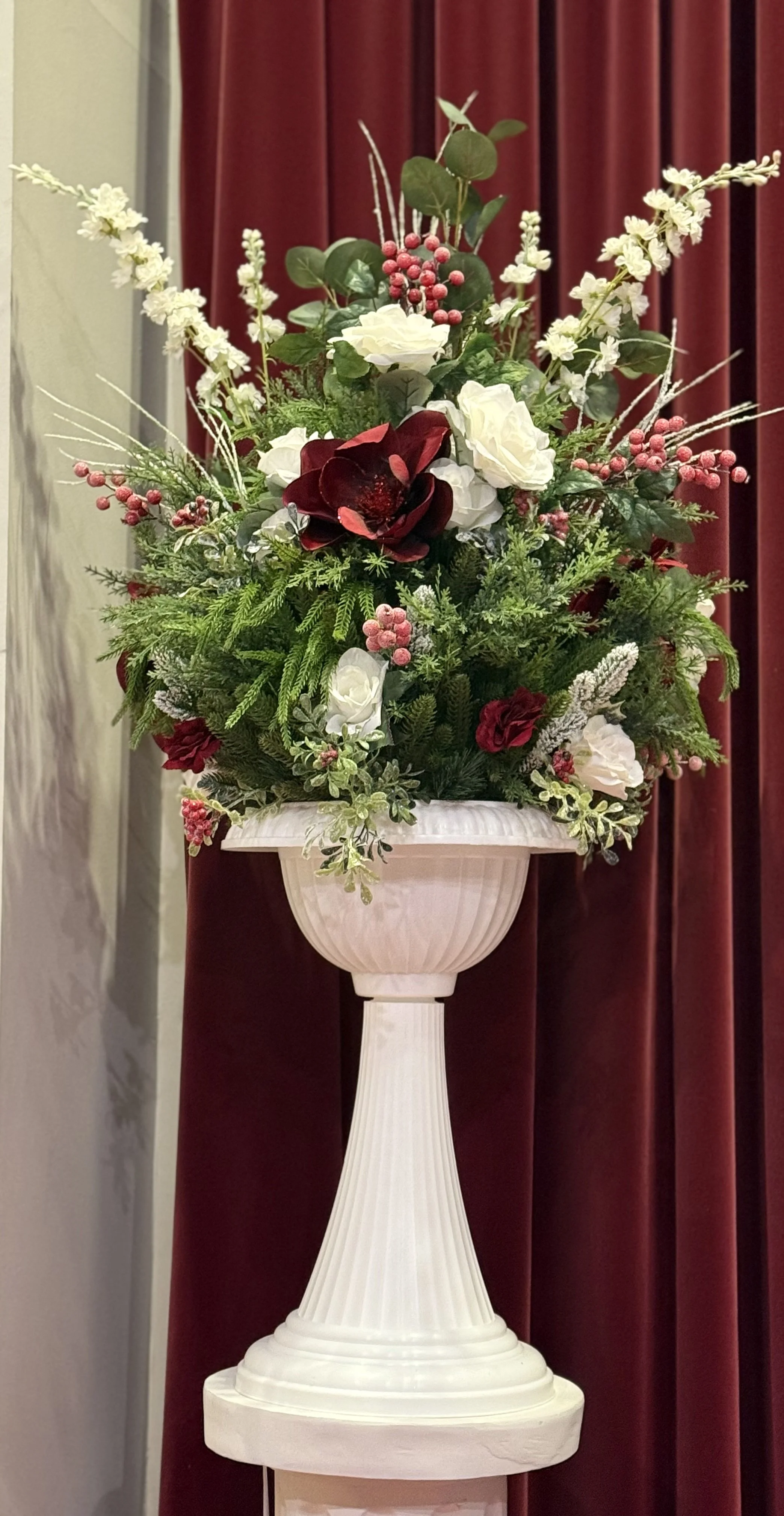A holiday floral arrangement with white and red flowers and green foliage in a white urn.