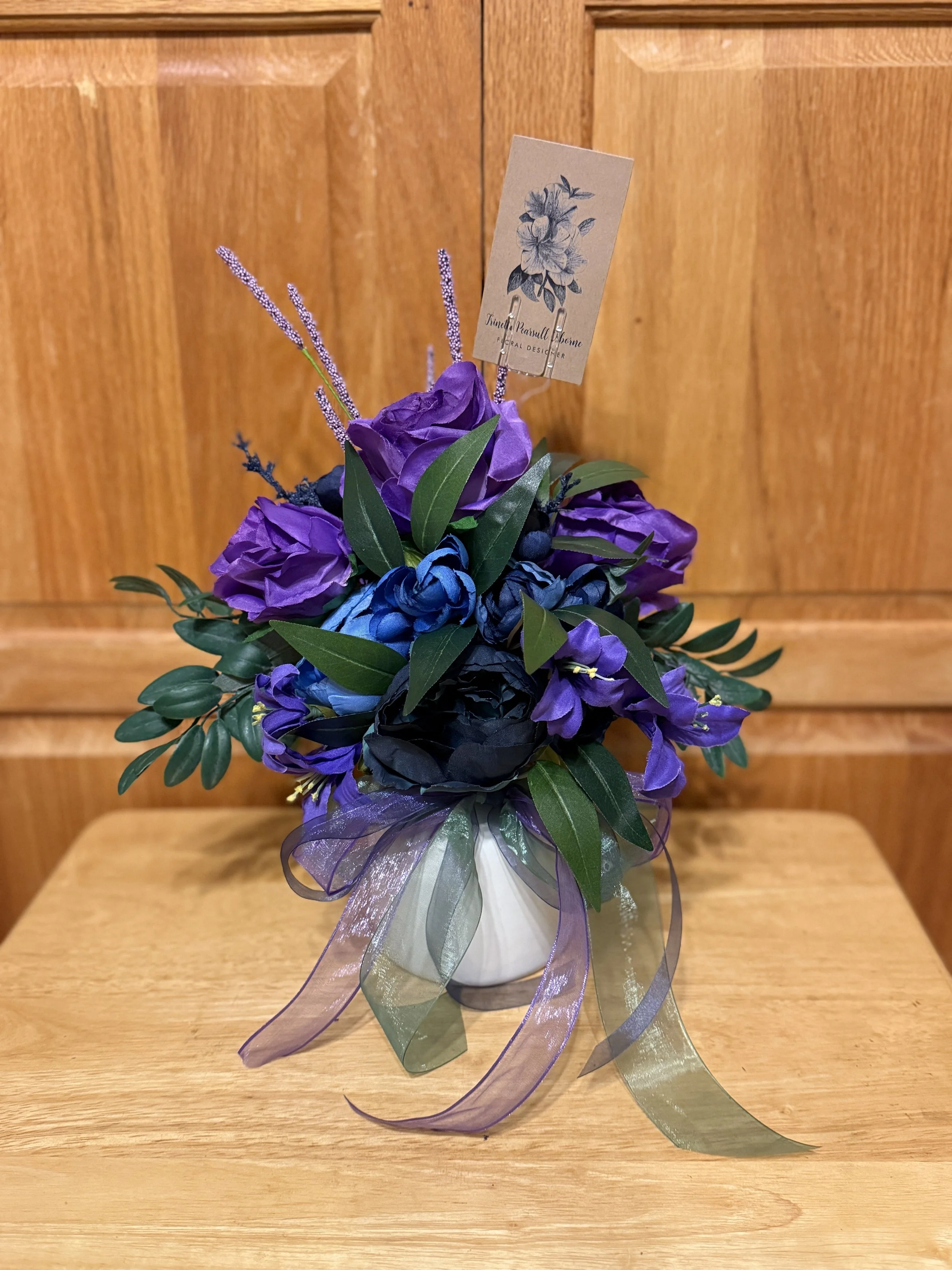 A purple and blue artificial flower arrangement in a white vase with a purple and green ribbon bow, placed on a wooden table in front of a wooden cabinet.