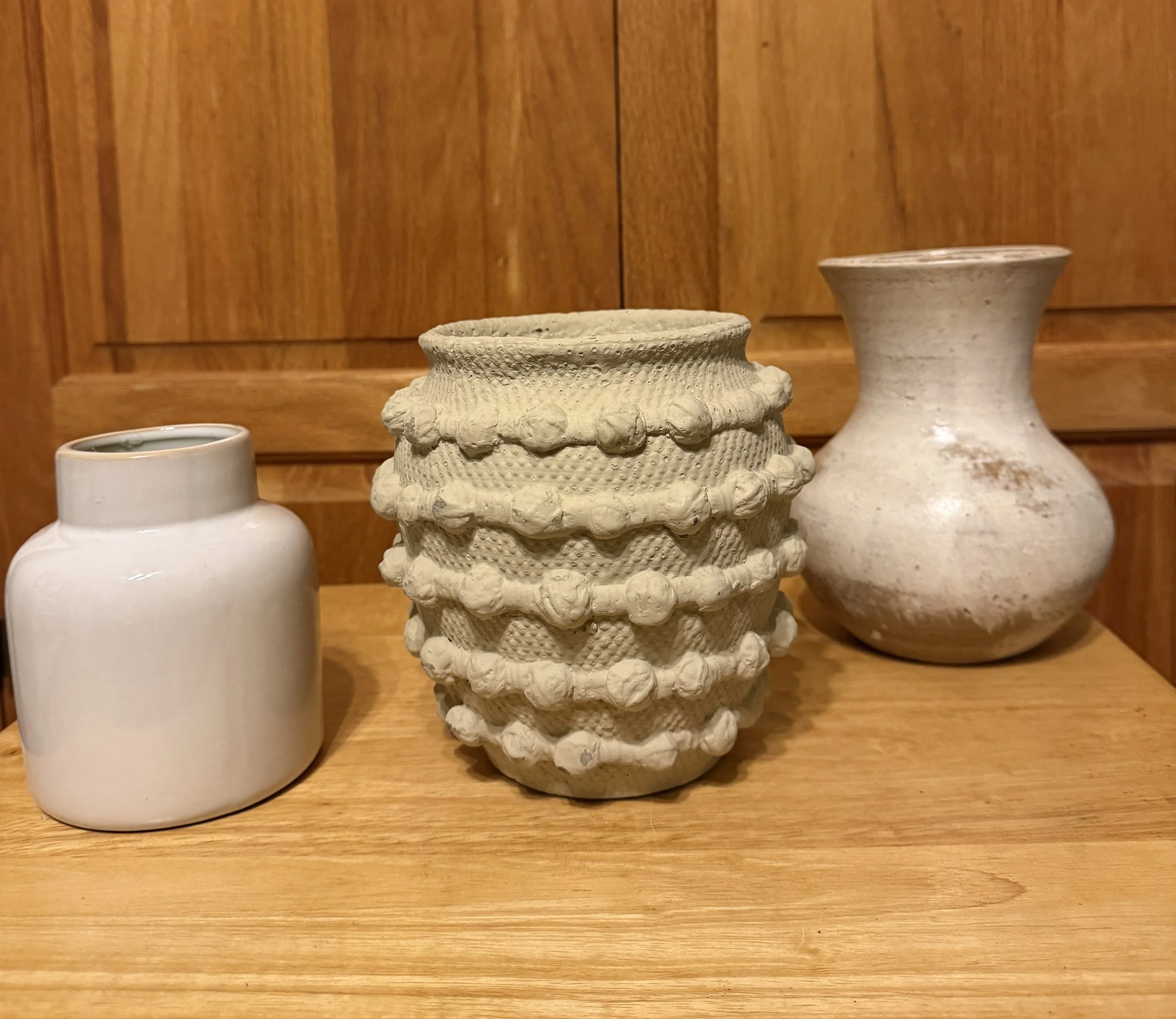 Three ceramic vases on a wooden surface against a wooden wall, with one small white vase on the left, a textured cream-colored vase with bobbles in the middle, and a larger, more rounded cream-colored vase on the right.
