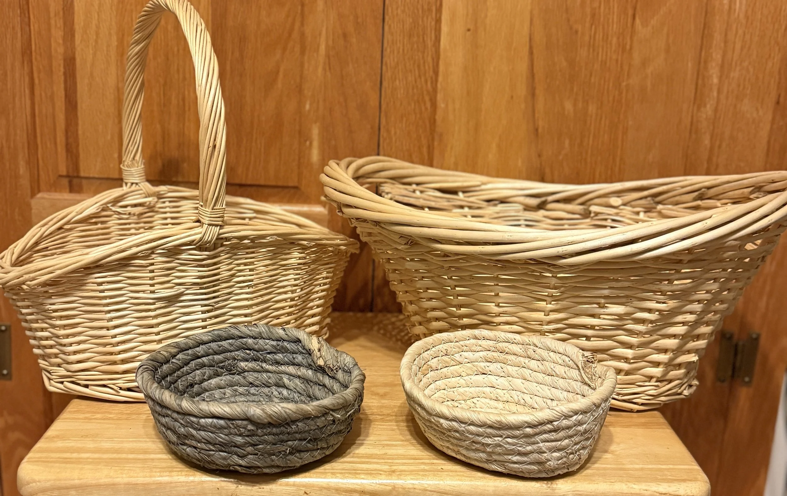 Four woven baskets, two large and two small, placed on a wooden surface against a wooden-paneled background.
