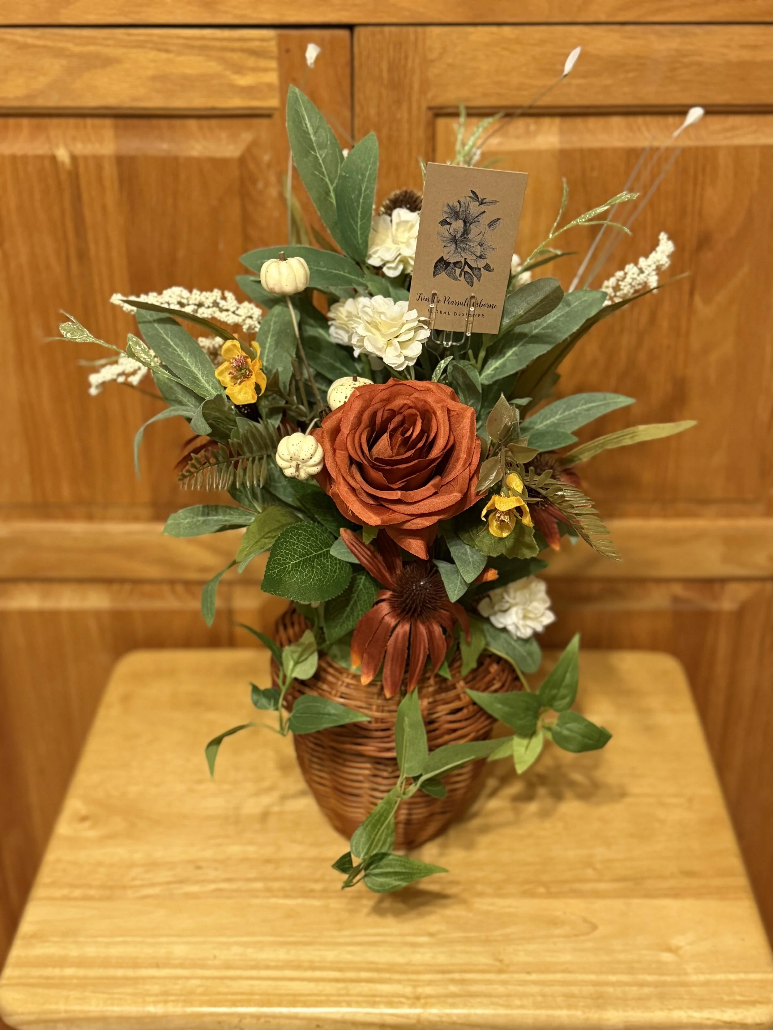 A floral arrangement in a wicker basket with orange, white, and yellow flowers, and various green leaves, placed on a wooden table against a wooden wall background.