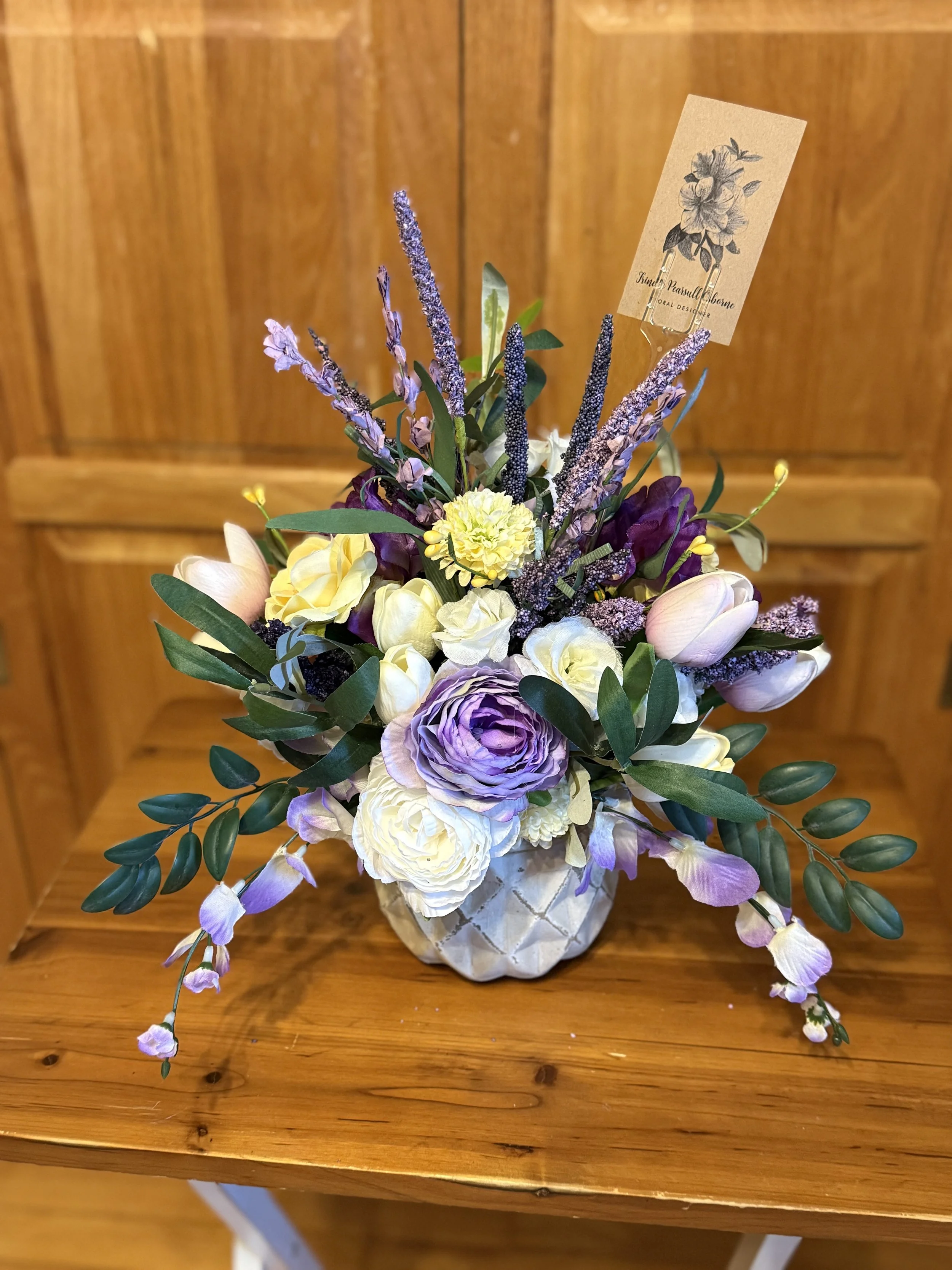 A floral arrangement in a decorative white ceramic pot, featuring purple, white, and yellow flowers with greenery, placed on a wooden surface against a wooden background.