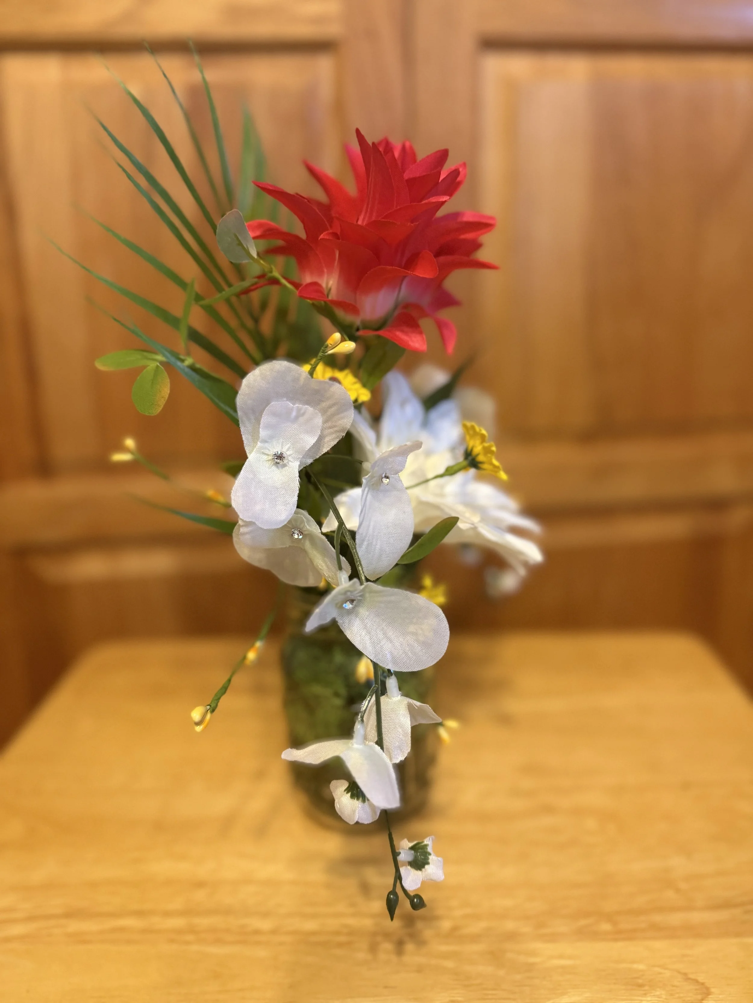 A flower arrangement in a vase with a red flower, white orchids, yellow fillers, and green leaves on a wooden table in front of a wooden background.