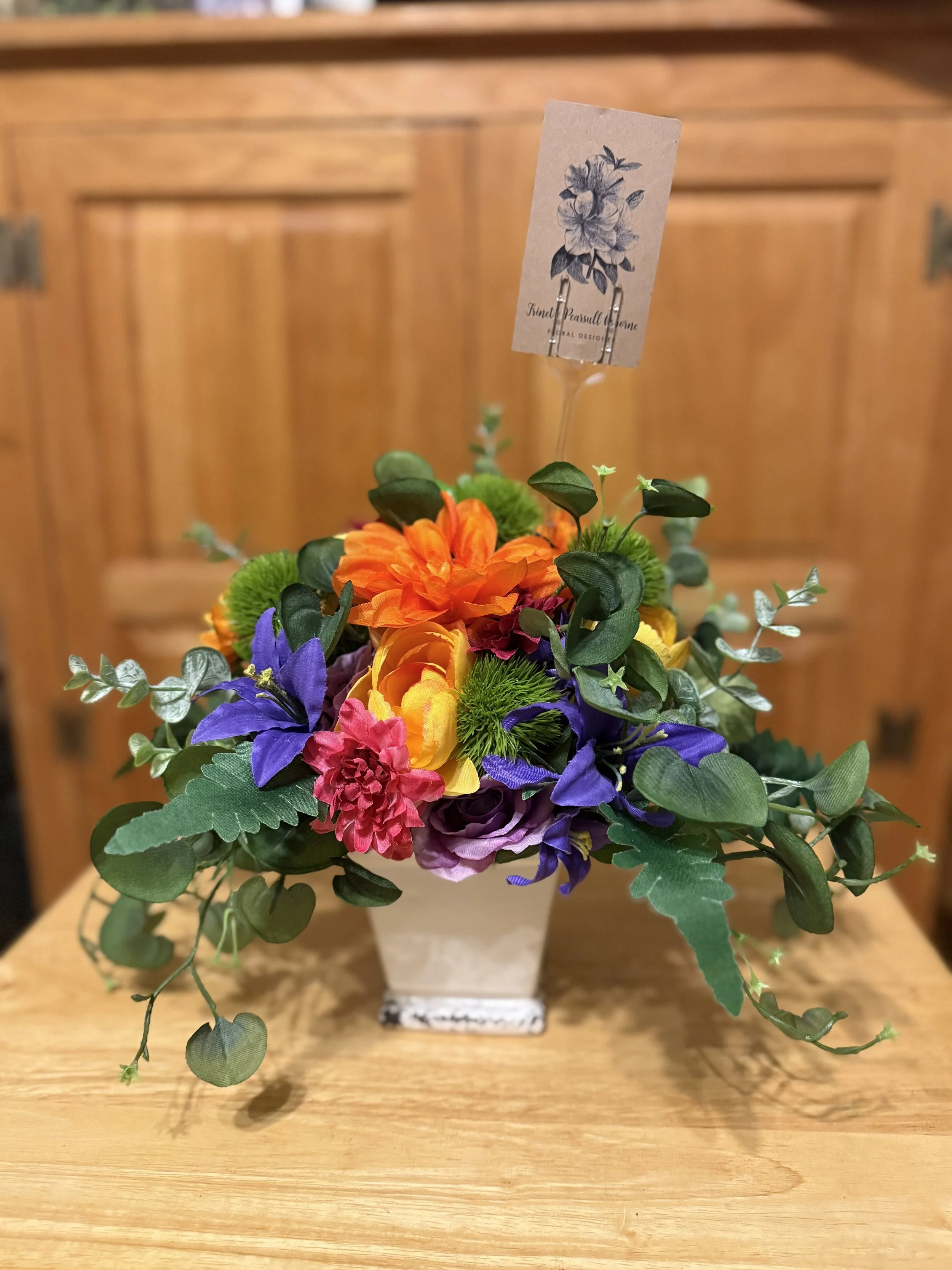 Colorful artificial flower arrangement in a square beige vase on a wooden surface, with a label attached reading 'Final Persuade Flower'.