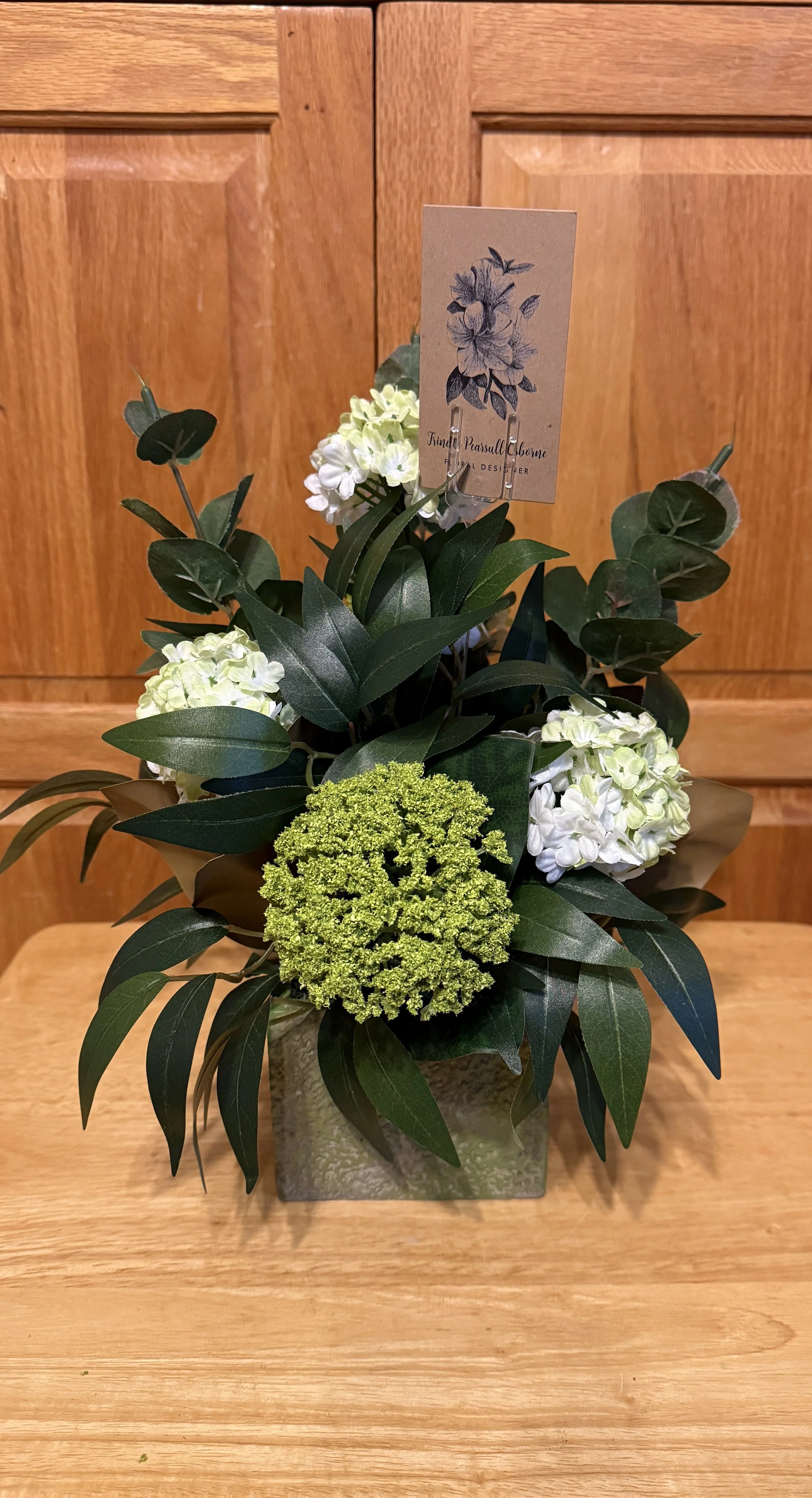 A floral arrangement with snow ball flowers, a light green flower, and green leaves in a square ceramic vase.