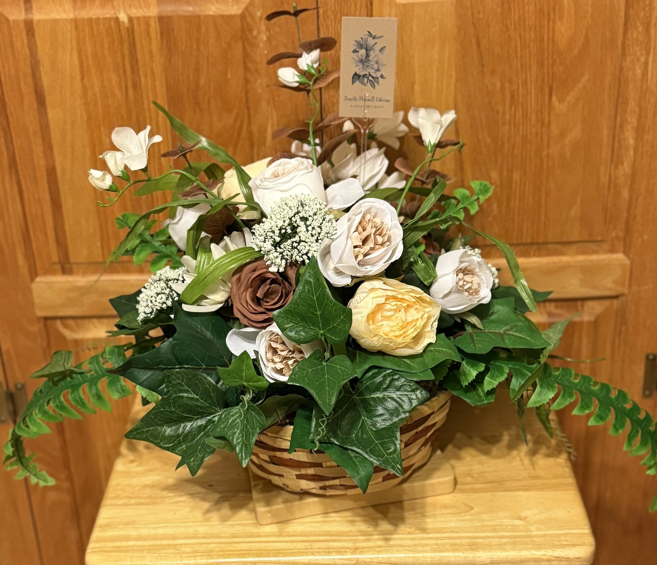 A basket of mixed flowers including white, cream, and brown roses, white filler flowers, and green foliage, set on a wooden surface.