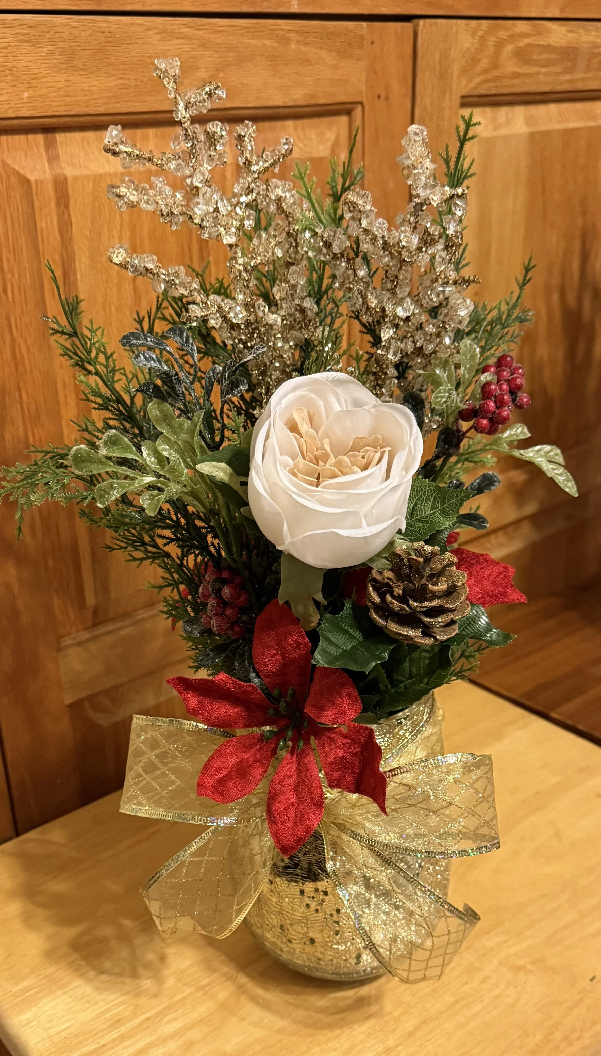 Holiday floral arrangement in a golden decorative vase, featuring a white rose, pinecones, red berries, greenery, and gold accents, tied with a gold ribbon.