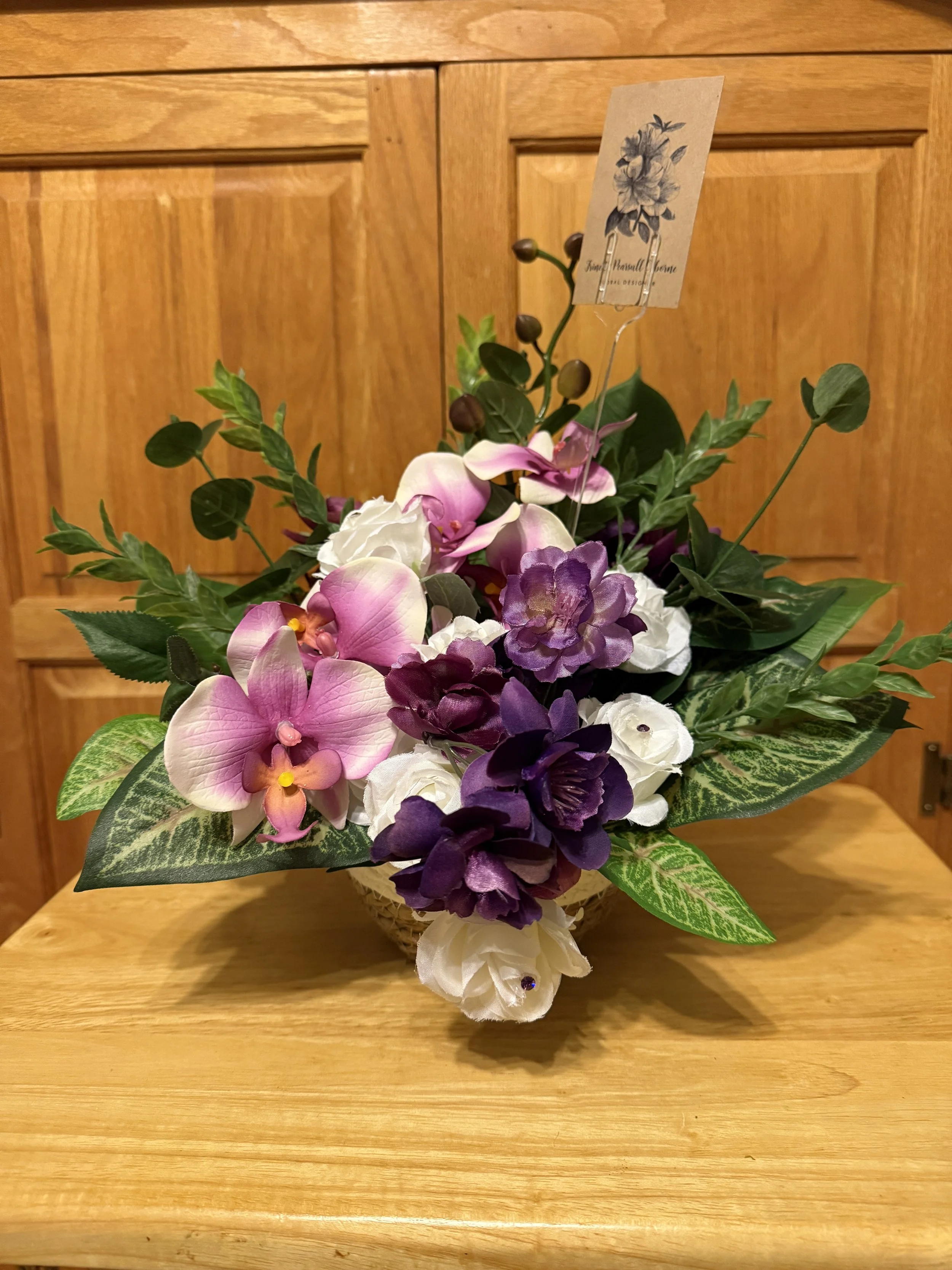 Arrangement of artificial flowers including orchids, roses, and greenery in a basket on a wooden table.