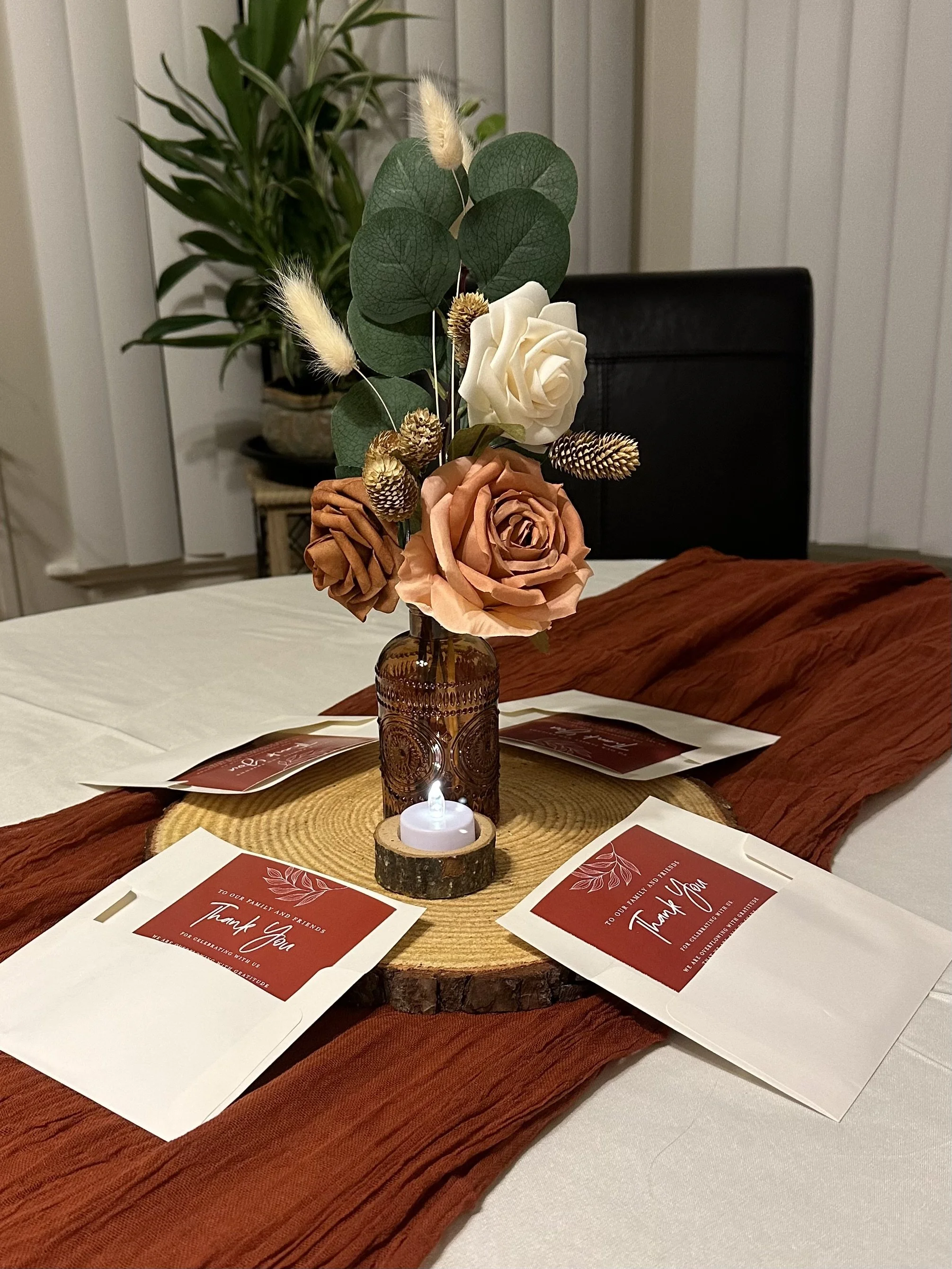 A table centerpiece featuring a floral arrangement of roses and greenery in a brown glass vase on a wooden slab, with a small tealight candle, surrounded by thank you cards on a white tablecloth with a rust-colored table runner.