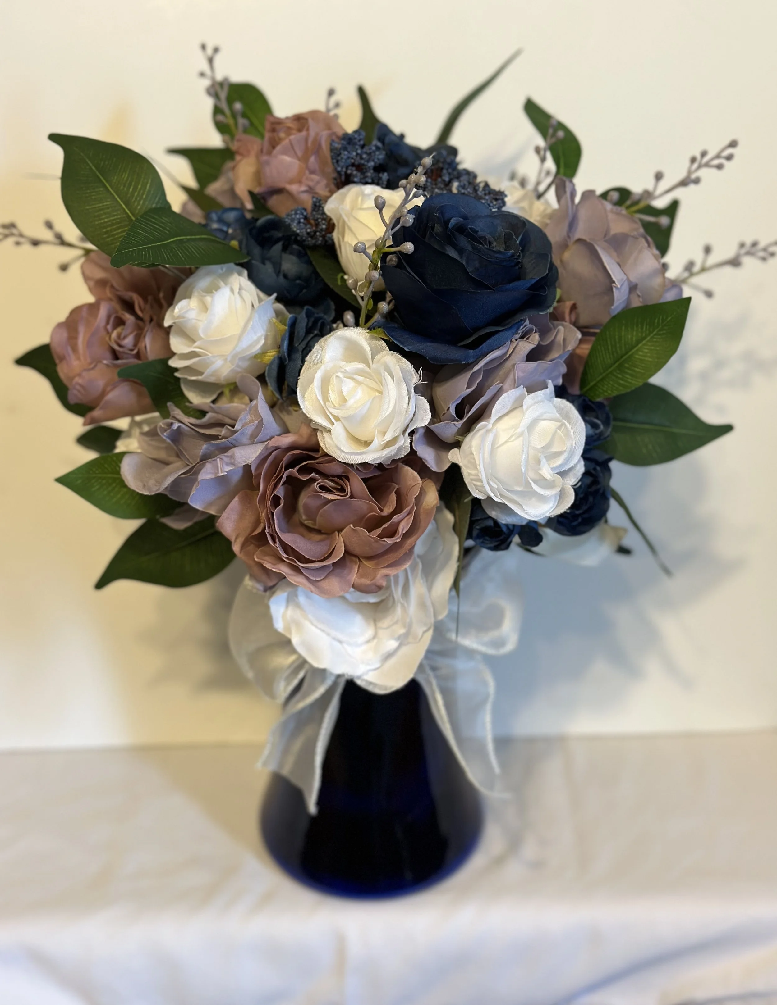 A bouquet of artificial roses in navy blue, white, and blush pink with green leaves, arranged in a black vase with a cream-colored ribbon tied around it.