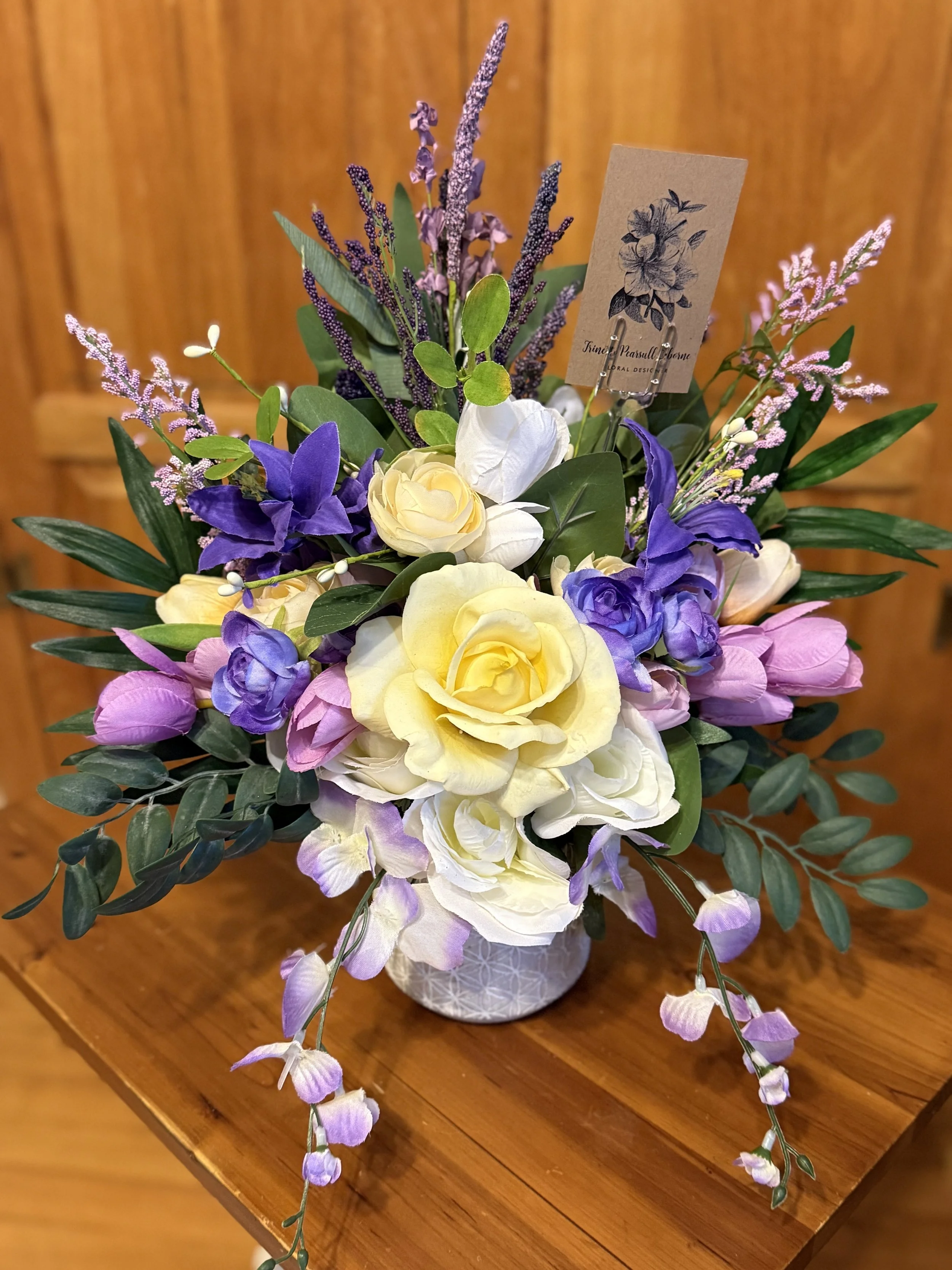 A floral arrangement featuring yellow roses, purple tulips, white freesias, lavender, and greenery in a white patterned vase placed on a wooden table.