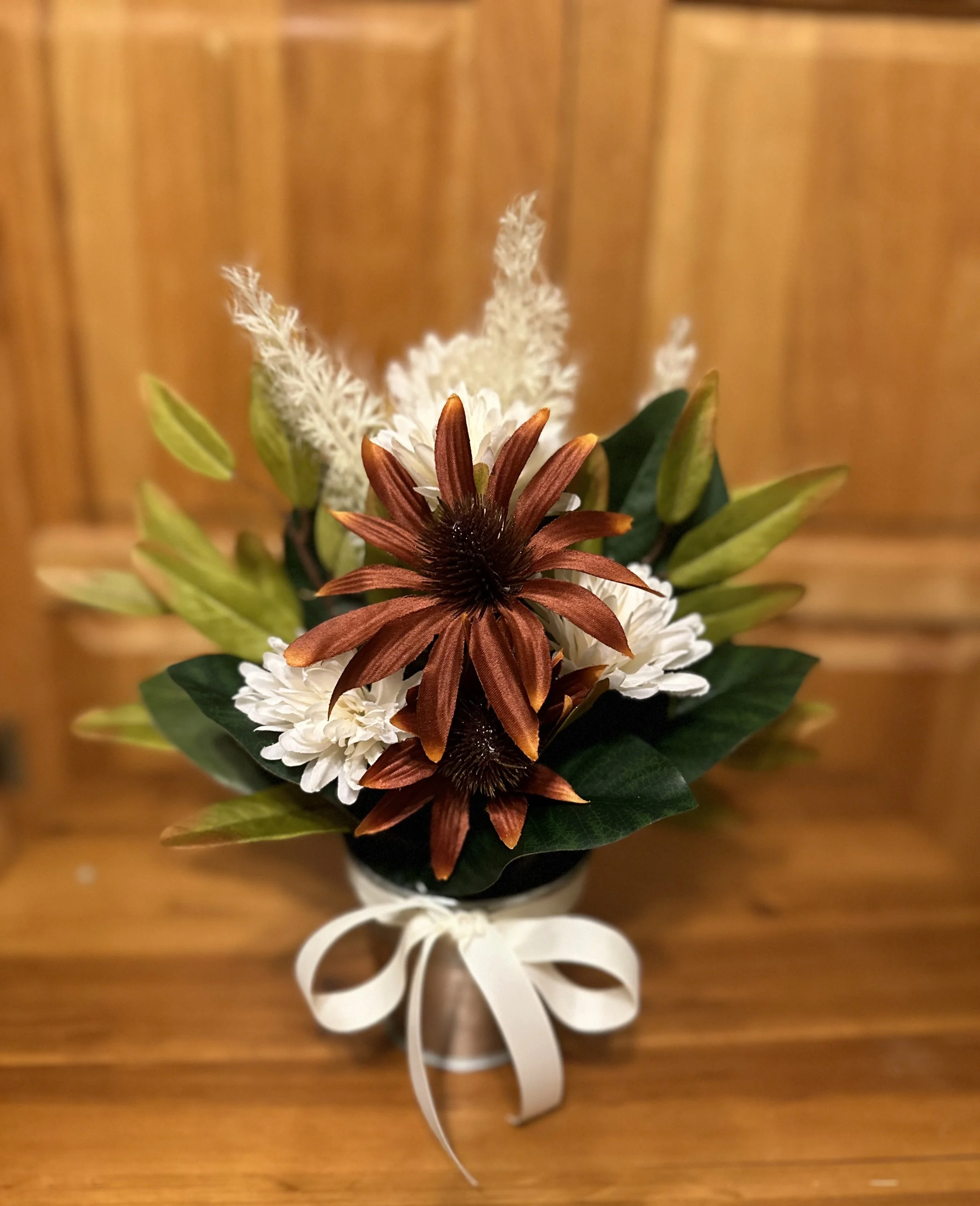 A floral arrangement with white, brown, and cream-colored flowers in a small vase with a white ribbon, set against a wooden background.
