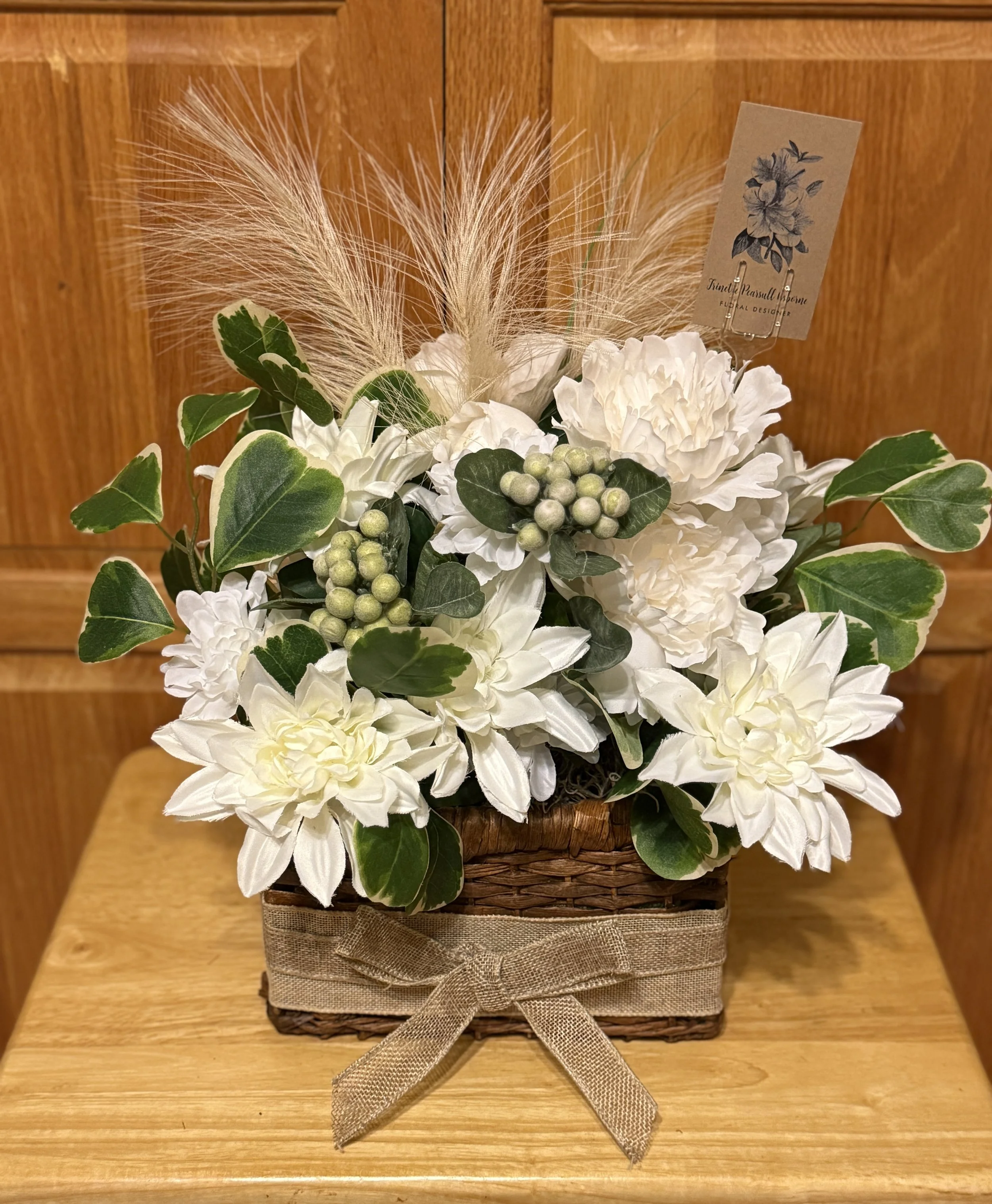 A floral arrangement in a woven basket decorated with a beige ribbon, featuring white flowers, green leaves, small green berries, and feathery accents, with a card attached.
