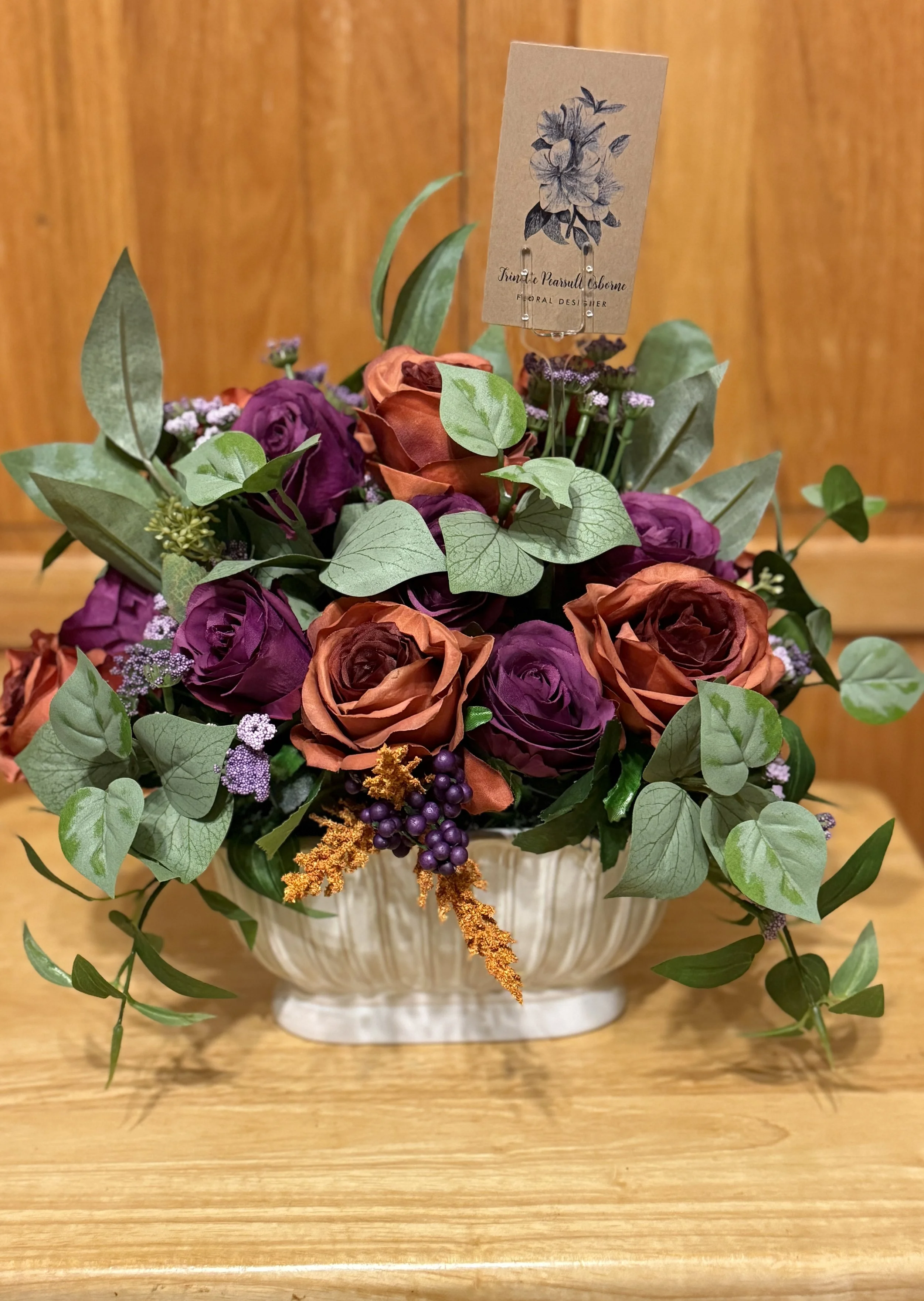 A floral arrangement with purple and rust-colored roses, green leaves, and small purple and white flowers in a cream-colored vase on a wooden surface.