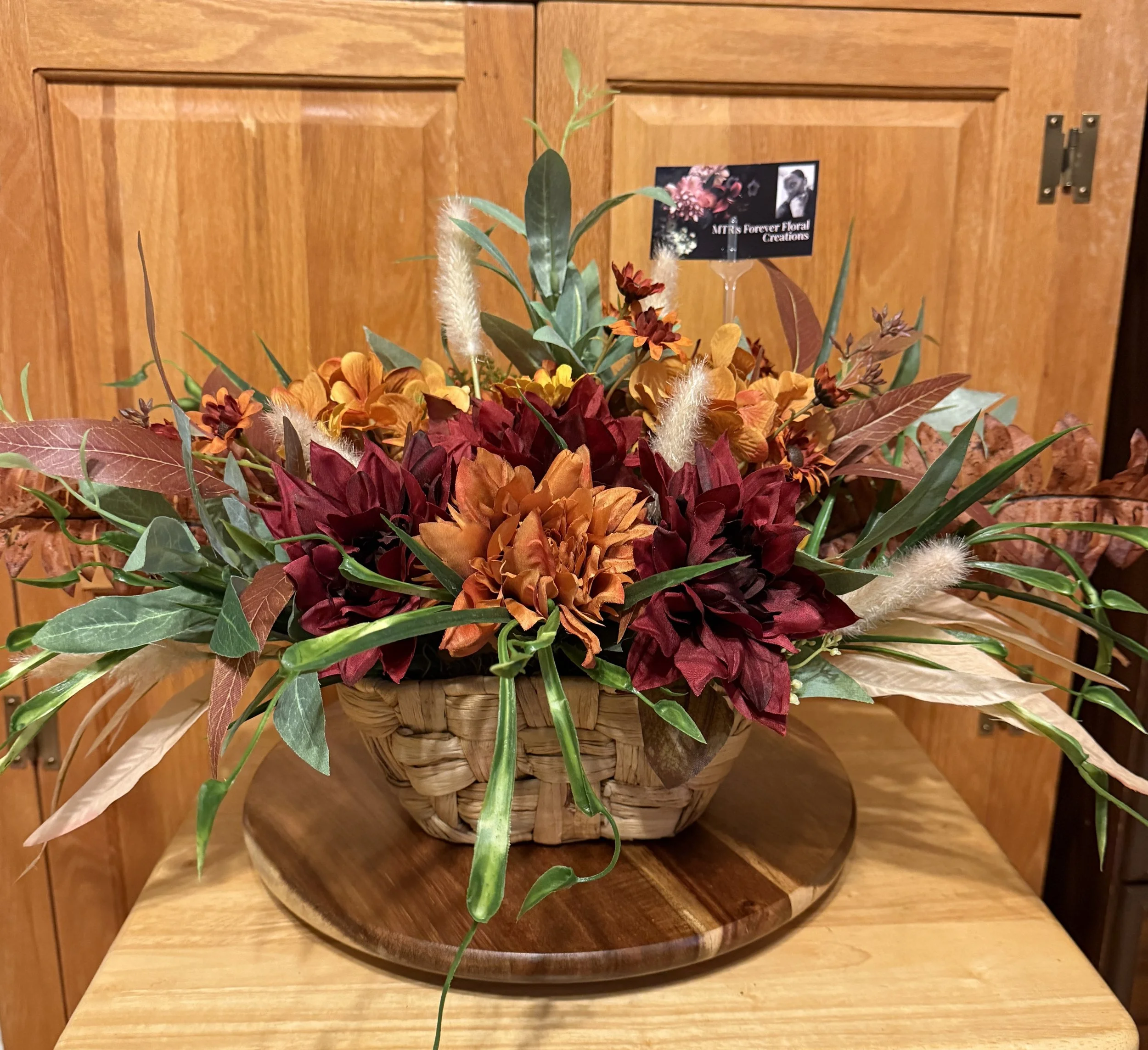 Autumn floral arrangement in a woven basket, containing orange, yellow, and burgundy flowers, with green, brown, and cream leaves, placed on a wooden table against a wooden cabinet background.