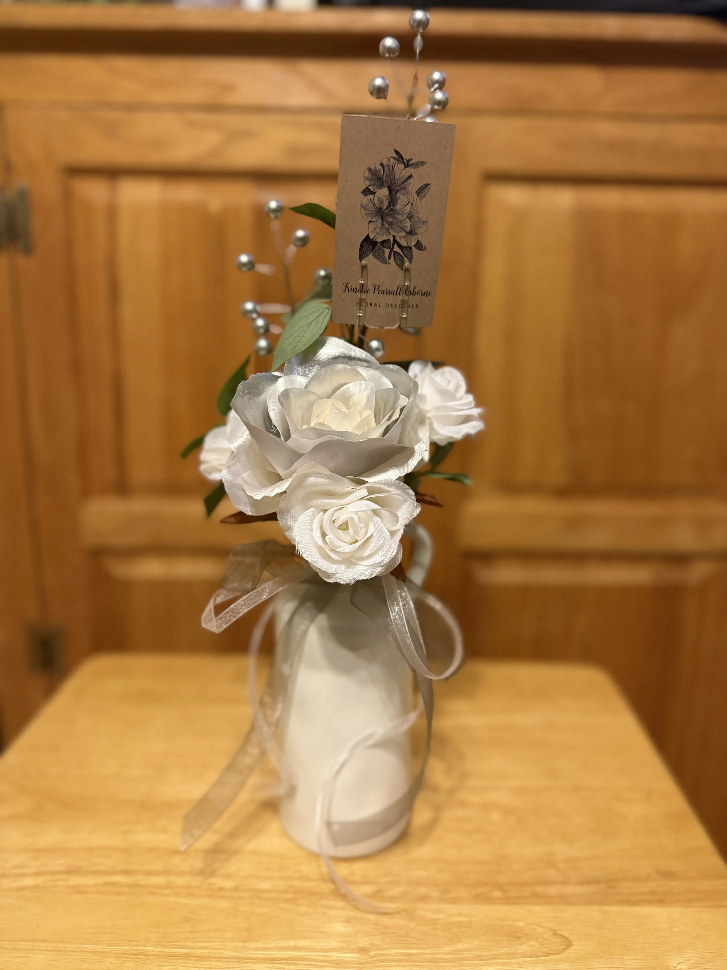 A white ceramic vase with a ribbon bow, holding white artificial roses, green leaves, and decorative silver pins, with a brown tag featuring a black floral illustration and text.