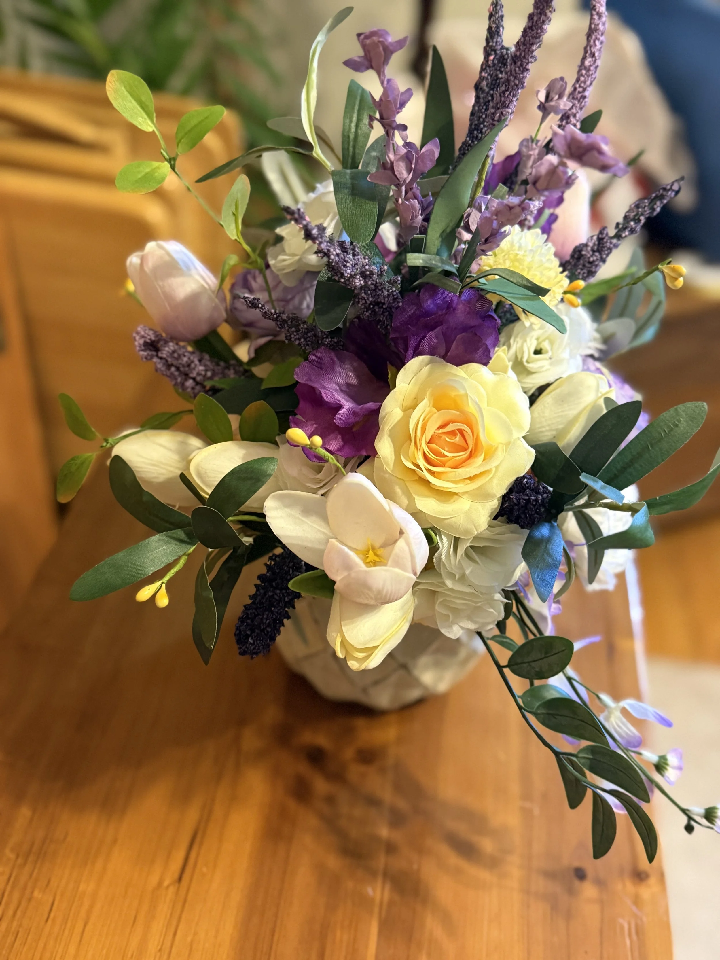 Colorful bouquet of yellow, purple, and white flowers with green leaves in a vase on a wooden table.