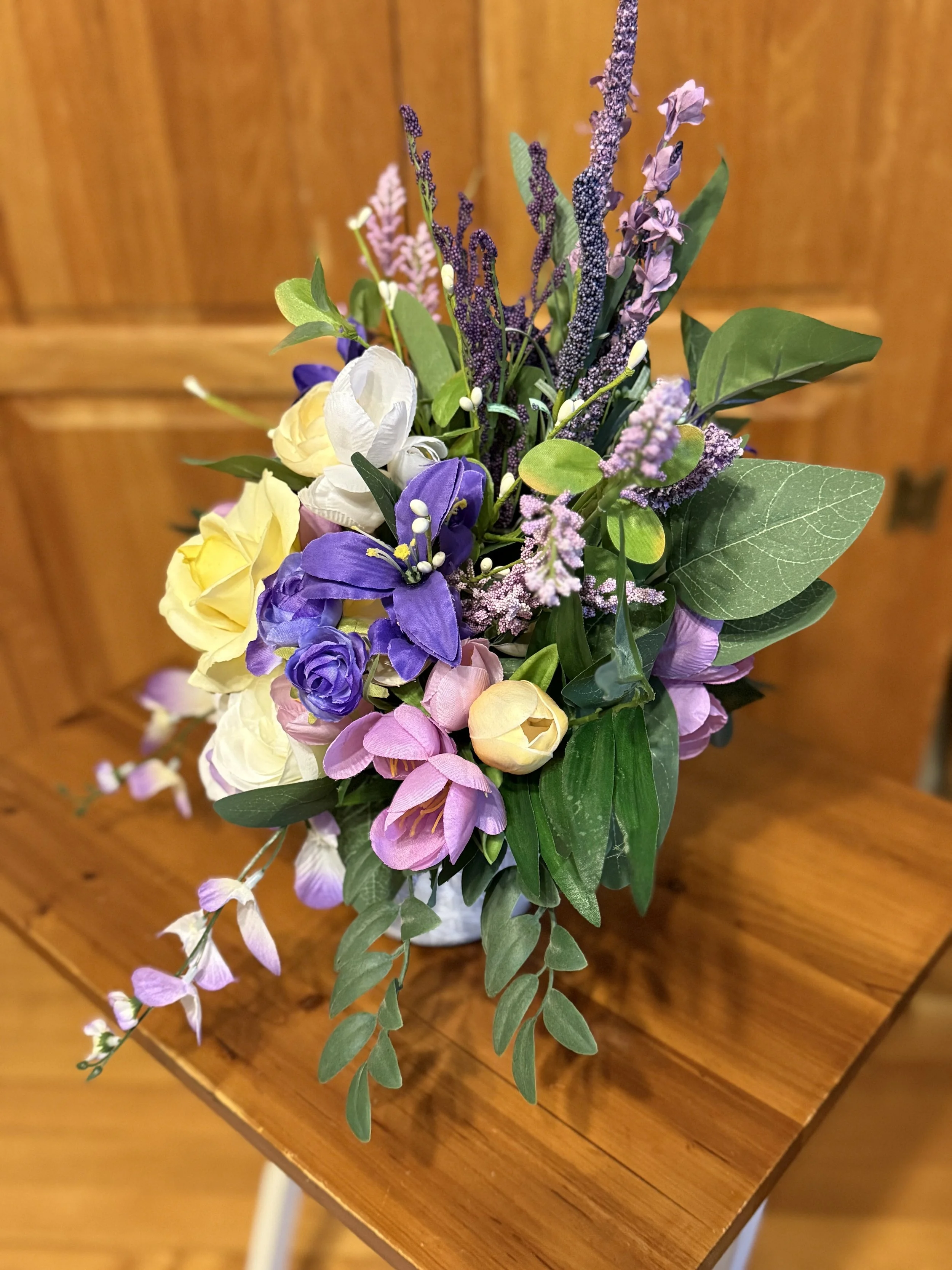 Colorful floral arrangement with purple, yellow, white, and pink flowers in a white vase on a wooden table.