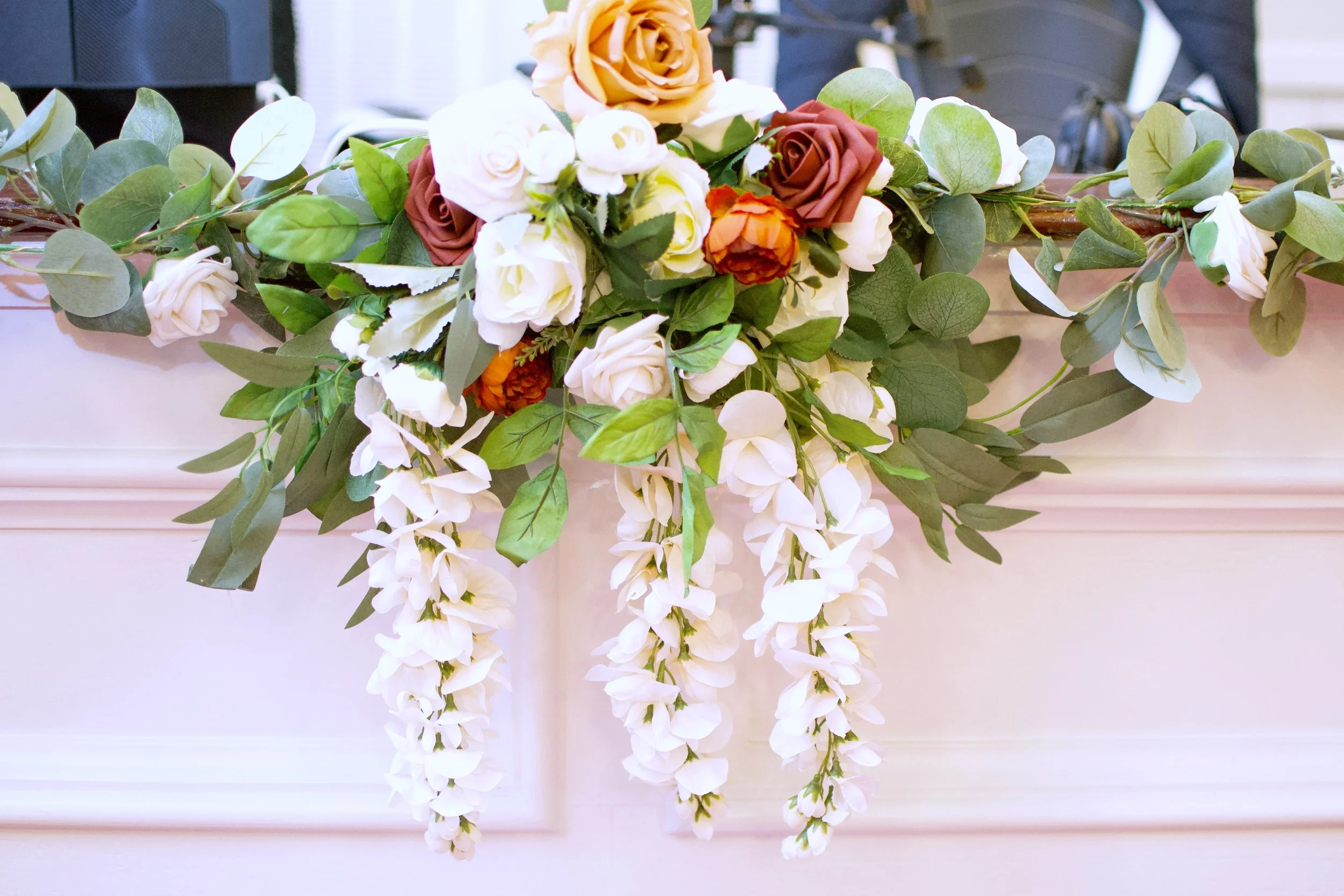 A floral swag with white, brown, and orange roses, cascading white wisteria, and green foliage.