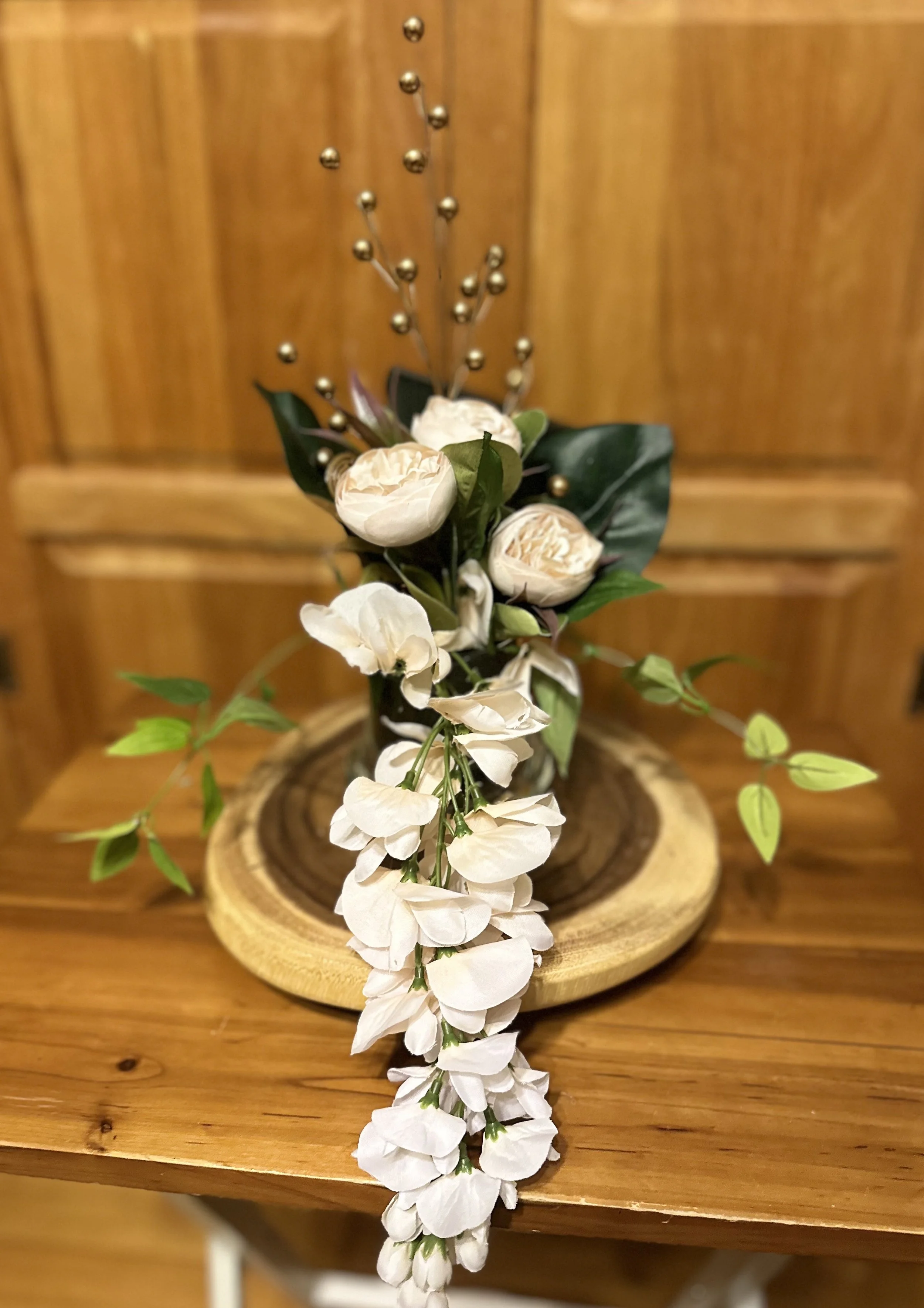 A floral arrangement with white roses, greenery, and decorative gold-colored branches on a wooden base.
