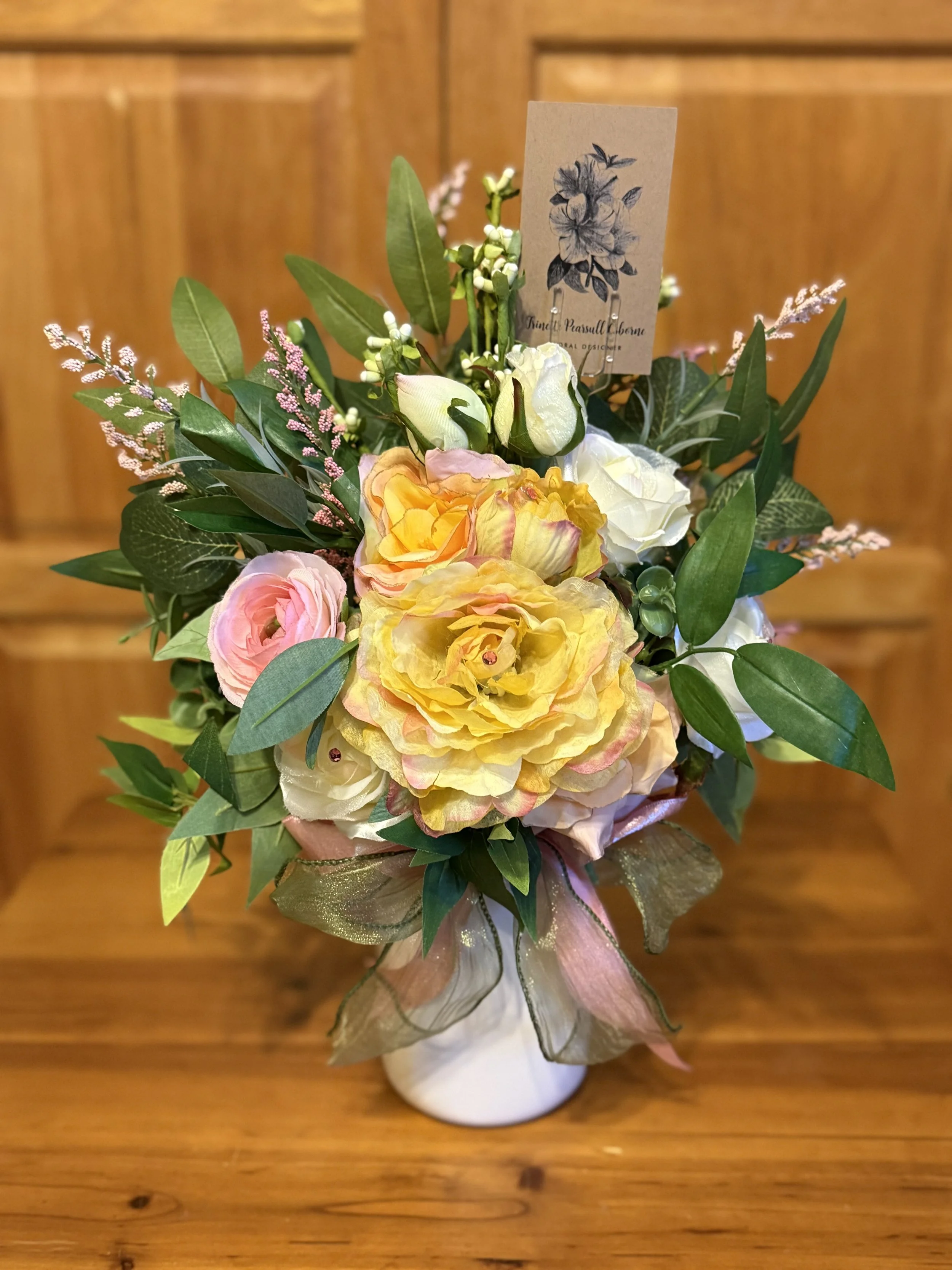 A colorful flower bouquet in a white mason jar vase on a wooden surface, composed of yellow, white, pink, and purple flowers with green leaves, and a decorative pink ribbon.
