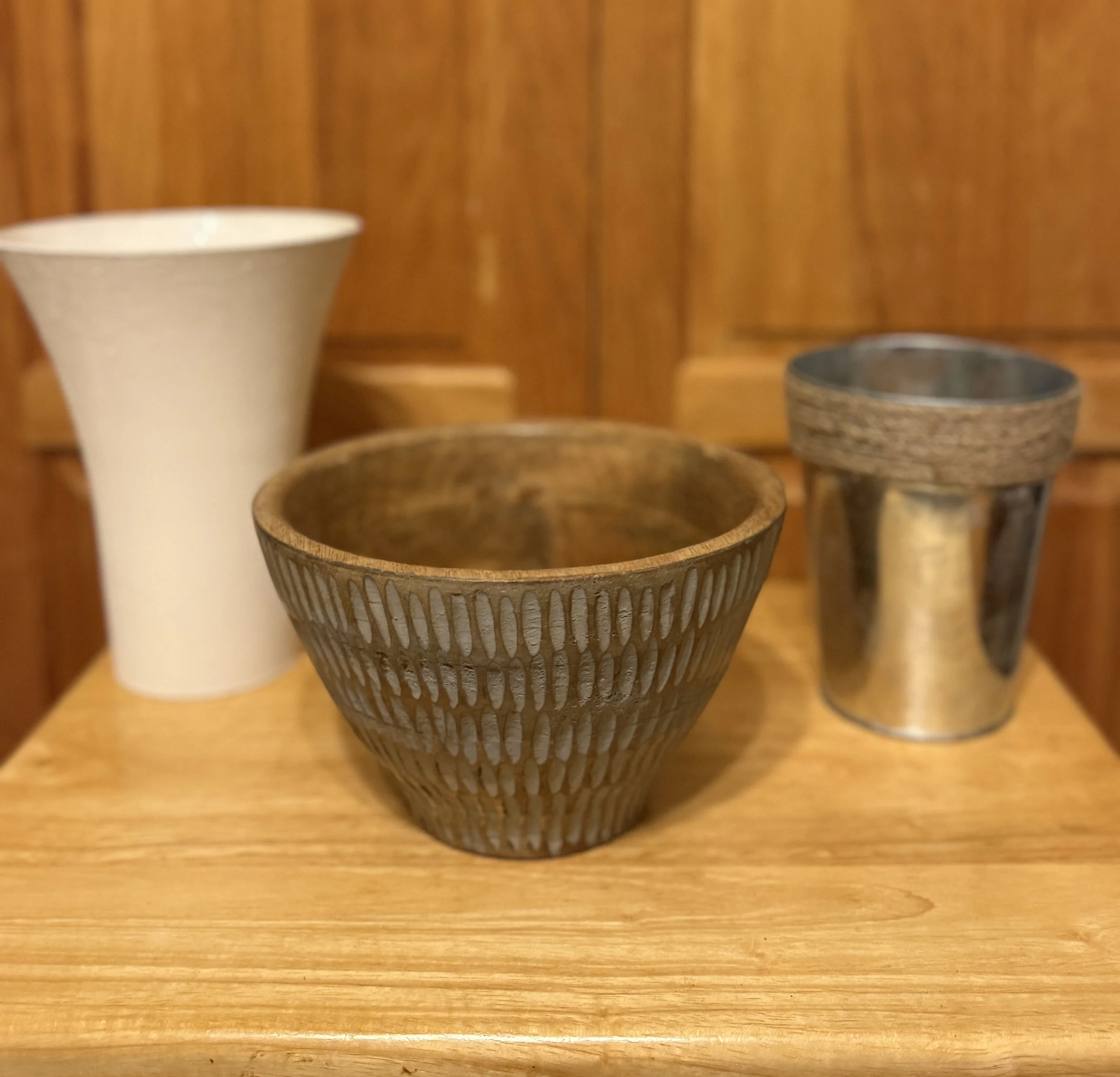 Three decorative bowls on a wooden surface in front of a wooden background, one white, one brown with vertical white streaks, and one metallic.