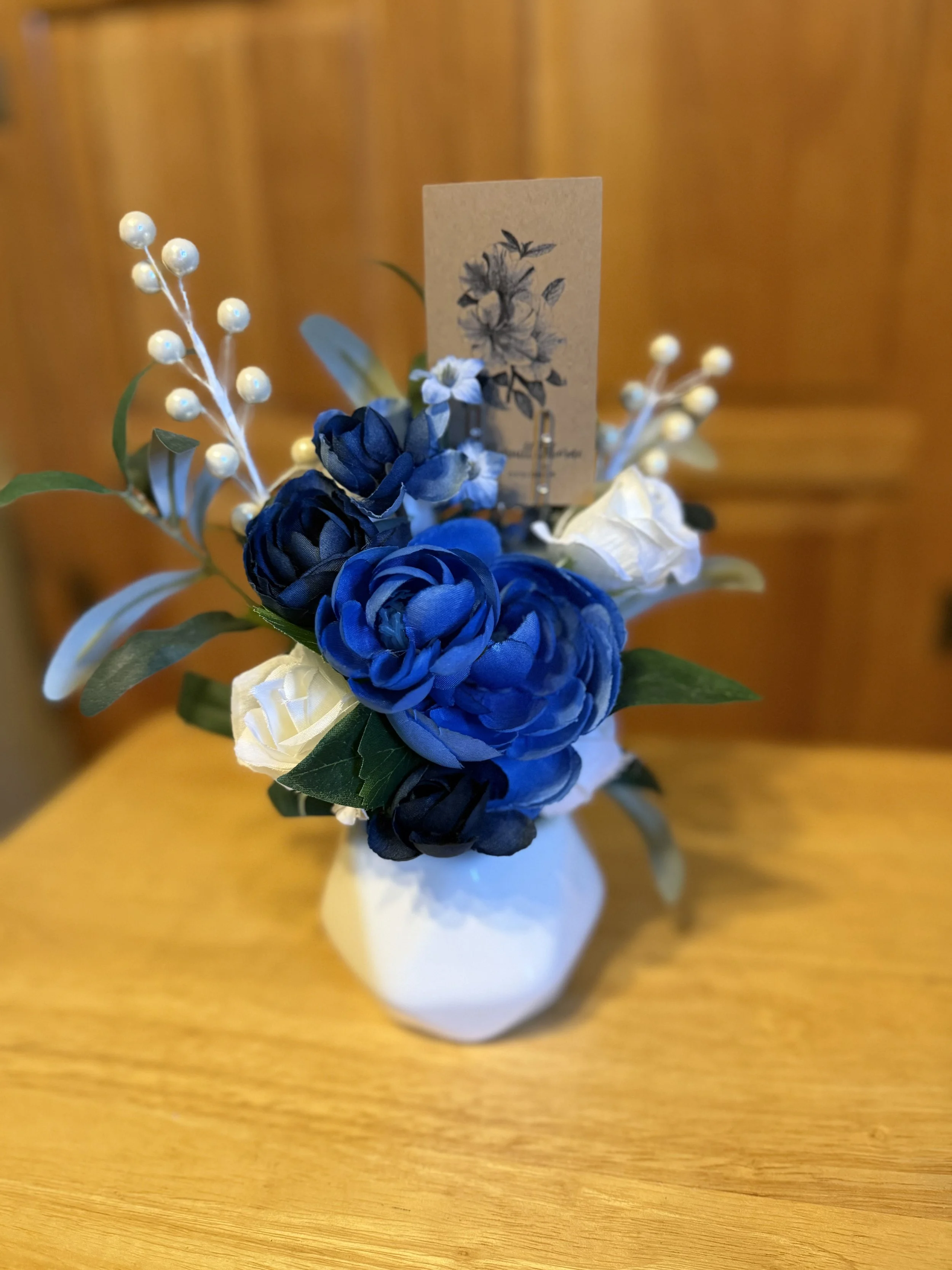 A small bouquet of blue and white artificial flowers in a white ceramic vase with a brown paper tag featuring a floral illustration, placed on a wooden surface.