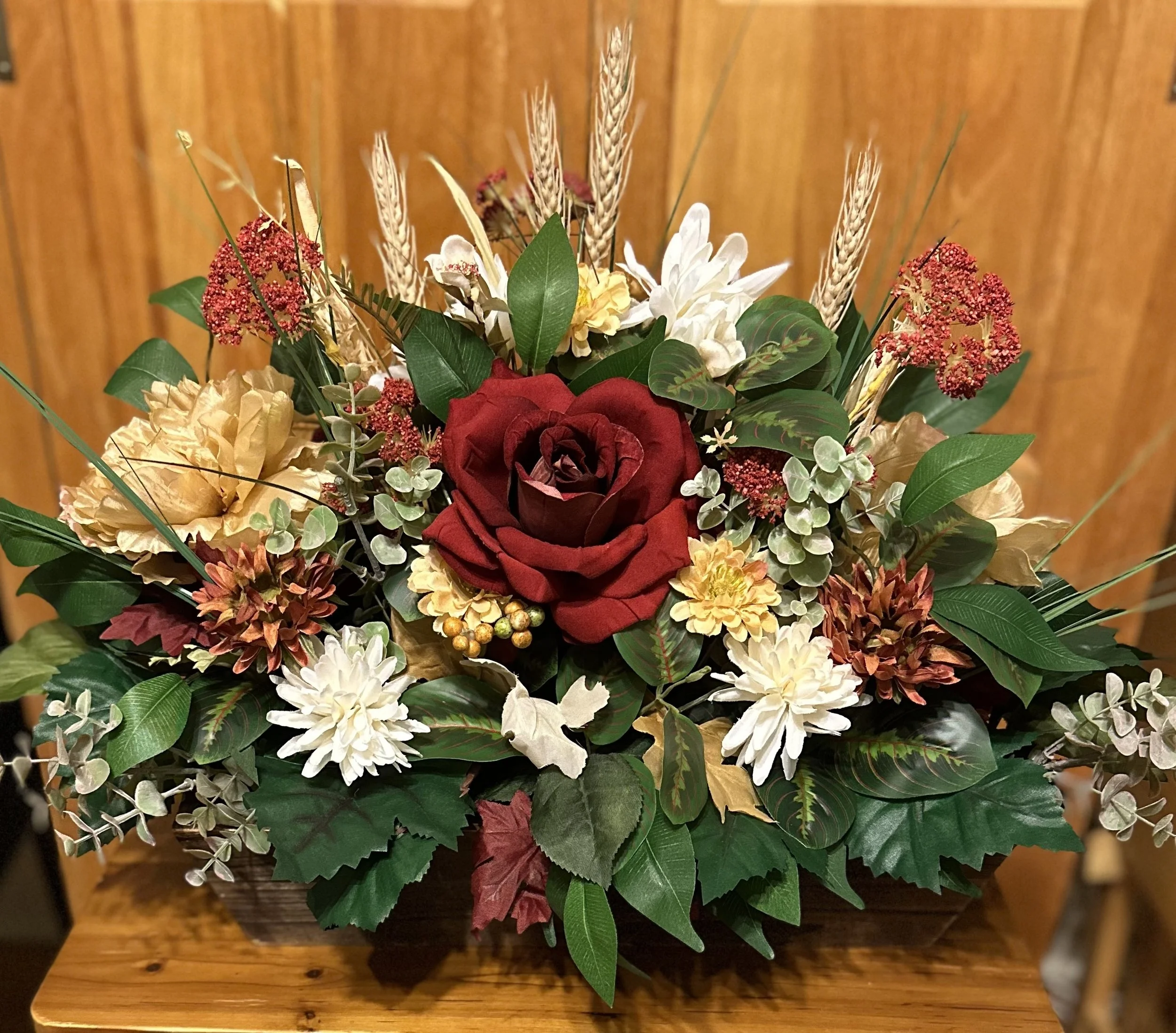A bouquet of flowers with a large red rose at the center, surrounded by white, yellow, and brown flowers, green leaves, and dried wheat stalks, arranged in a rectangular wooden container against a wooden background.