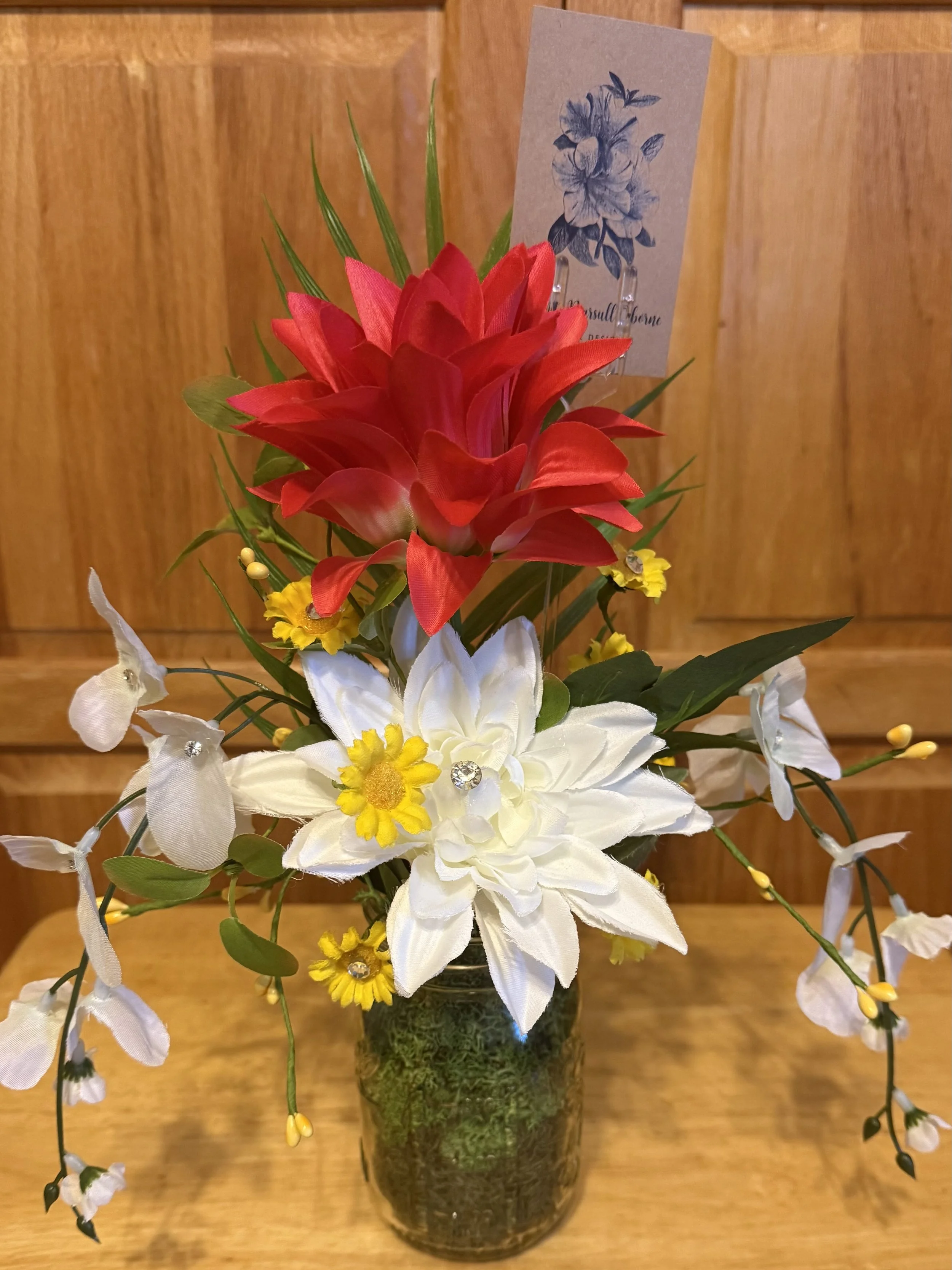 Flower arrangement in a glass vase with red, white, and yellow artificial flowers, and a thank you card with a blue floral illustration, set against a wooden background.