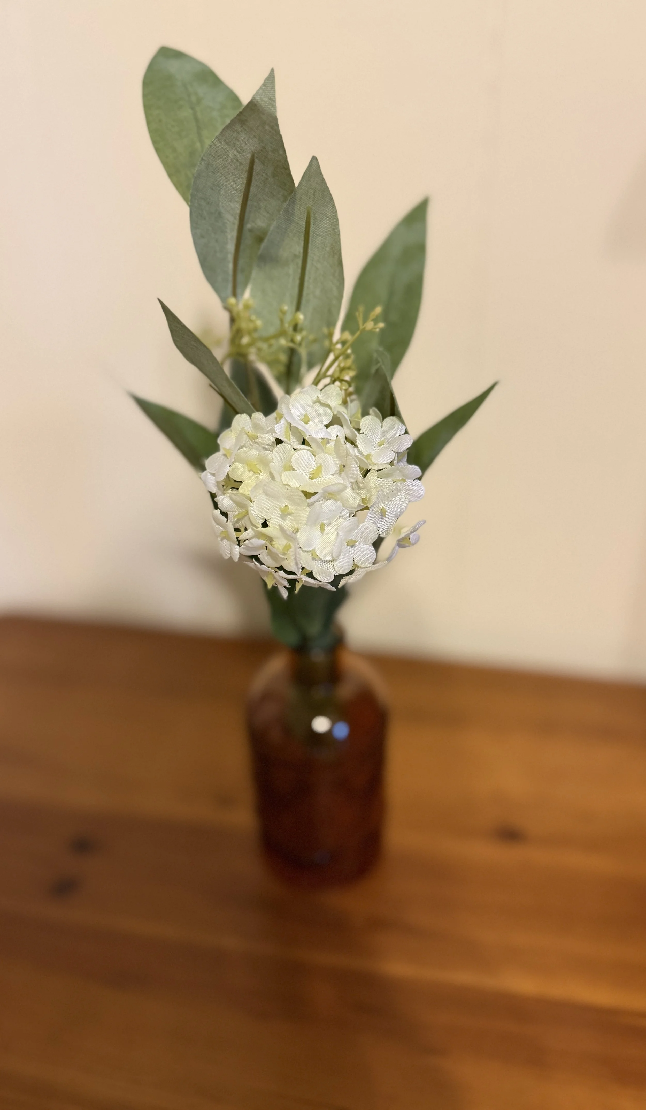 A small amber glass bottle with white snow ball flower and green leaves on a wooden surface.