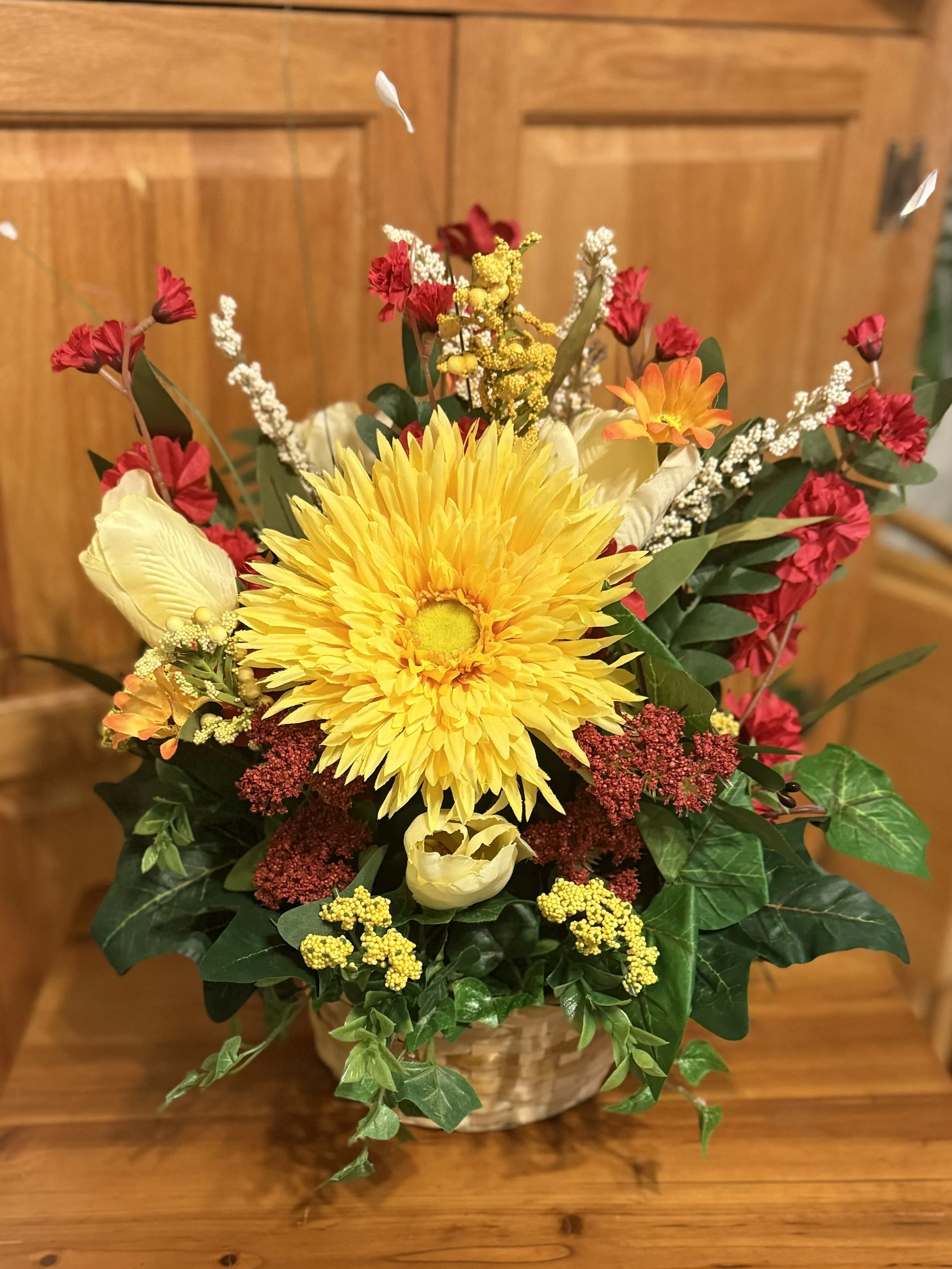 A colorful flower arrangement in a woven basket on a wooden table in front of a wooden cabinet, featuring a large yellow sunflower, red and white flowers, greenery, and small yellow and white accent flowers.