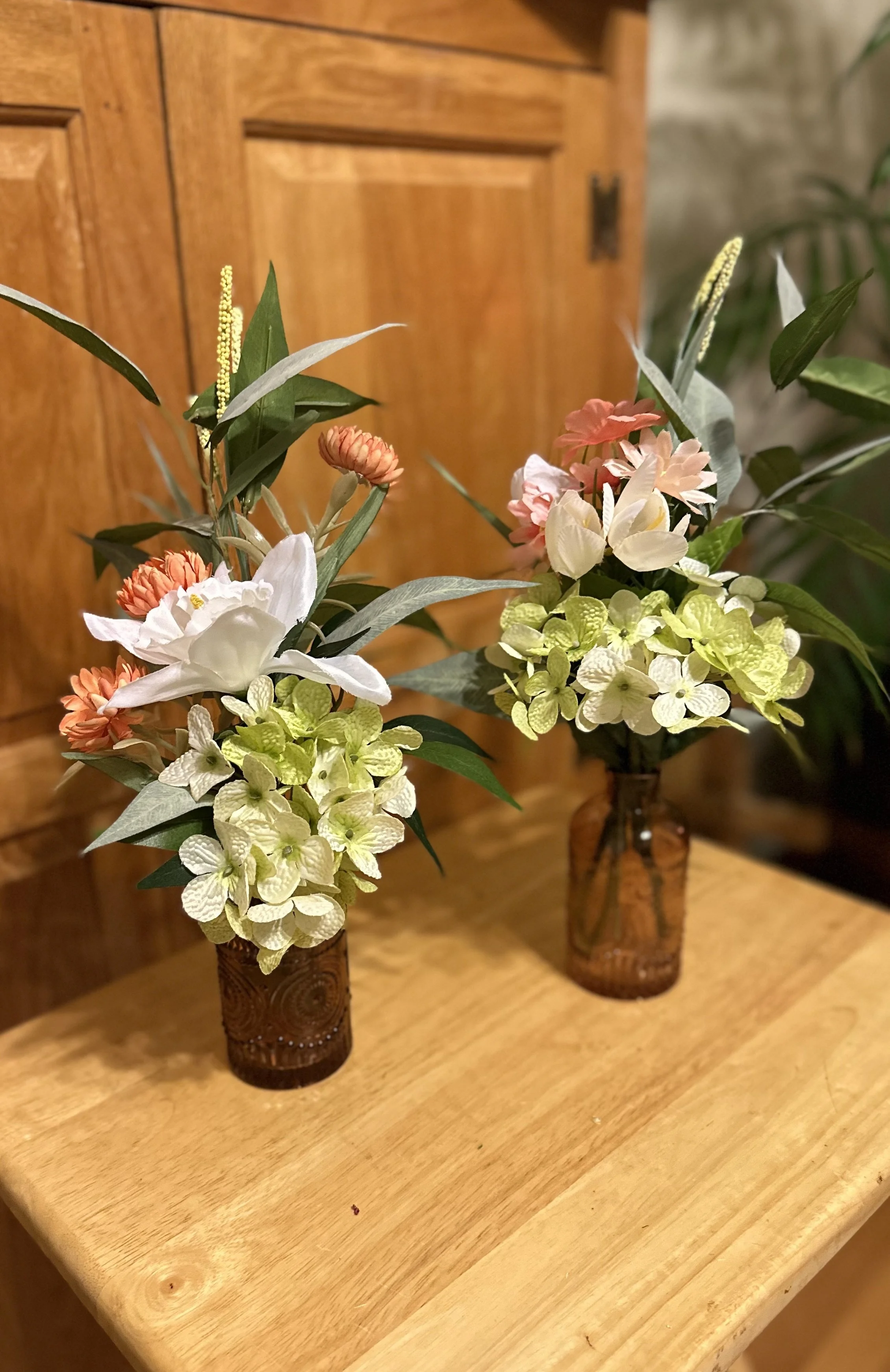 Two floral arrangements in brown glass vases with pink, white, and green flowers on a wooden surface against a wooden cabinet background.