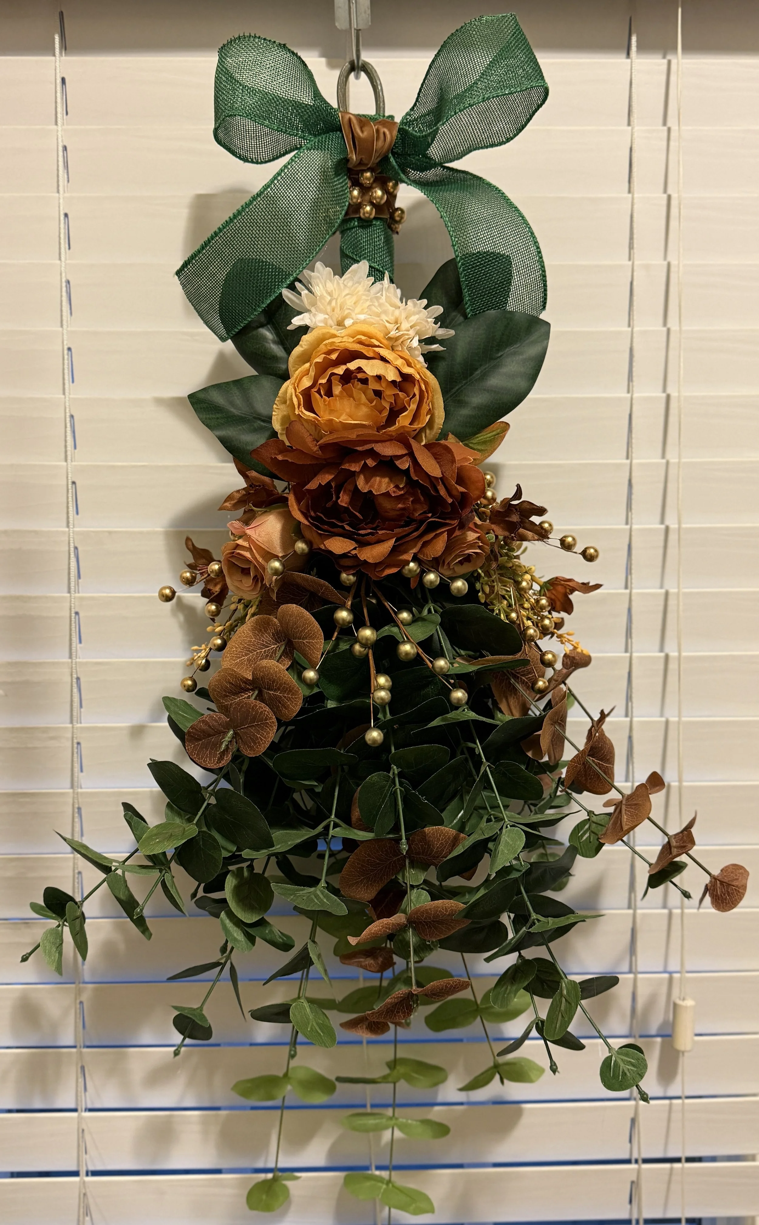Artificial floral decoration with green bow and gold beads hanging on white blinds.