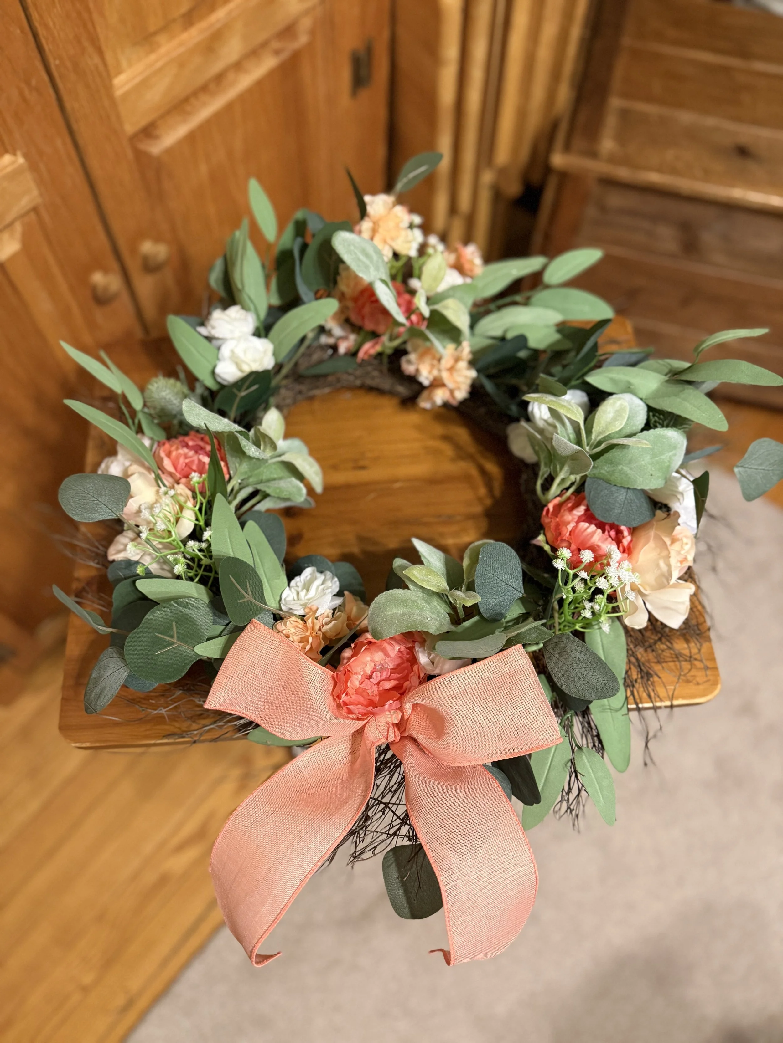 A decorative flower wreath with pink, white, and peach carnations, green leaves, and a pink ribbon bow, placed on a wooden surface.