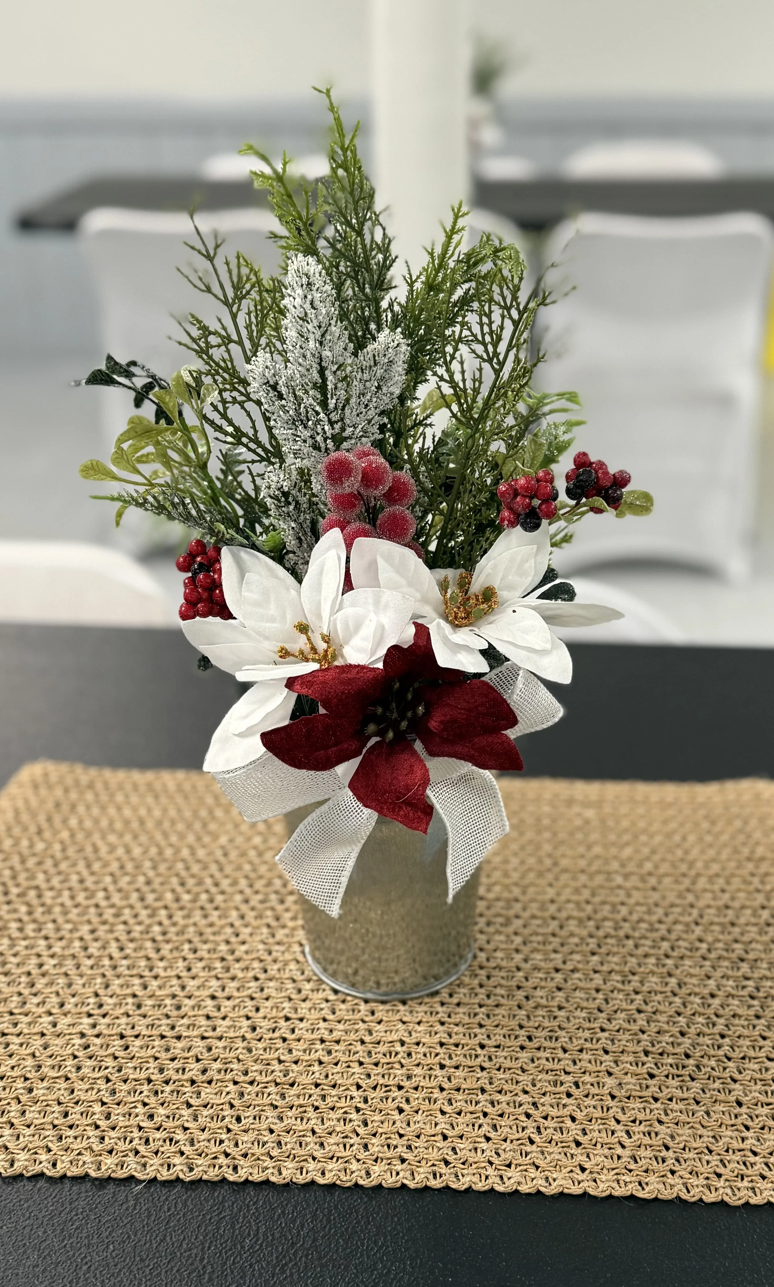 A festive holiday arrangement in a silver tin pot on a woven placemat, featuring white poinsettias, red berries, green foliage, and frosted accents.