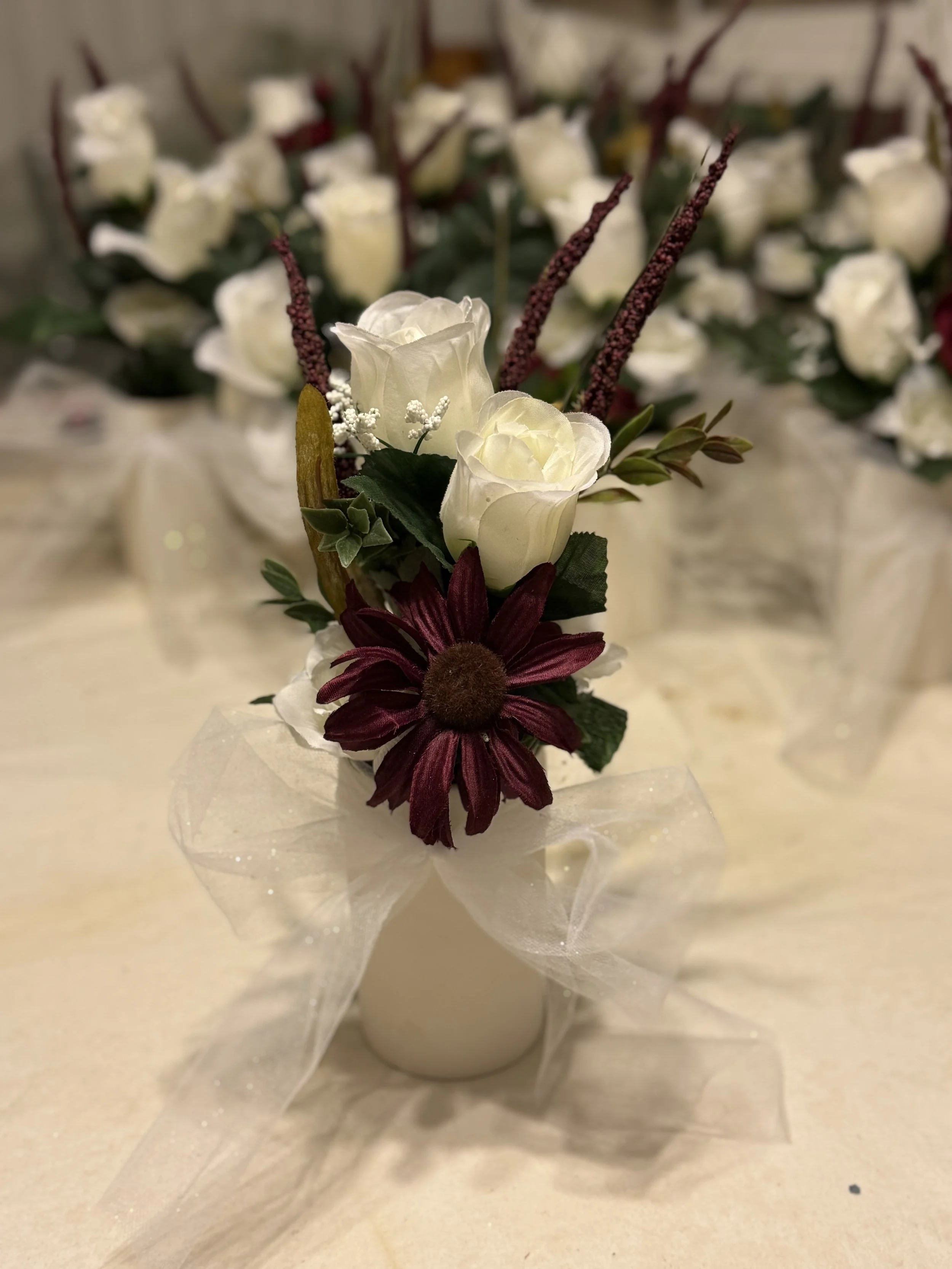 Small white flower arrangement with white roses, burgundy daisy, and greenery in a white vase, decorated with sheer white ribbon, on a table with a larger floral centerpiece in the background.