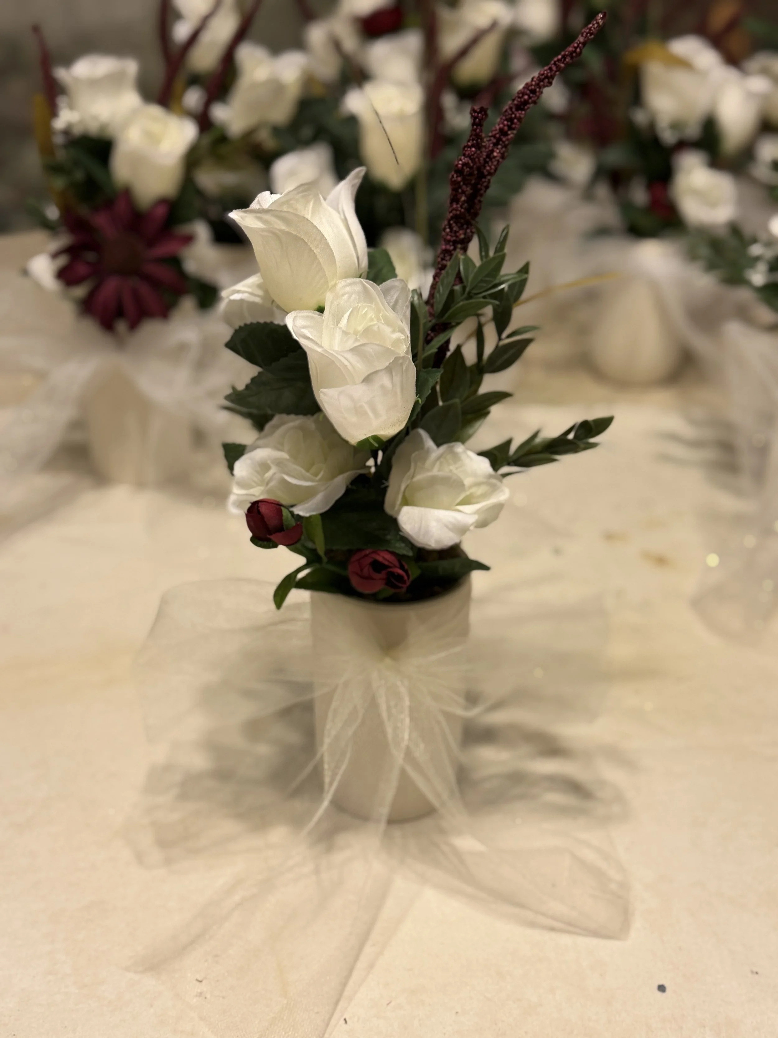 Small flower arrangement with white roses, small red flowers, and greenery in a white pot, decorated with a sheer ribbon bow, placed on a cream-colored tablecloth.