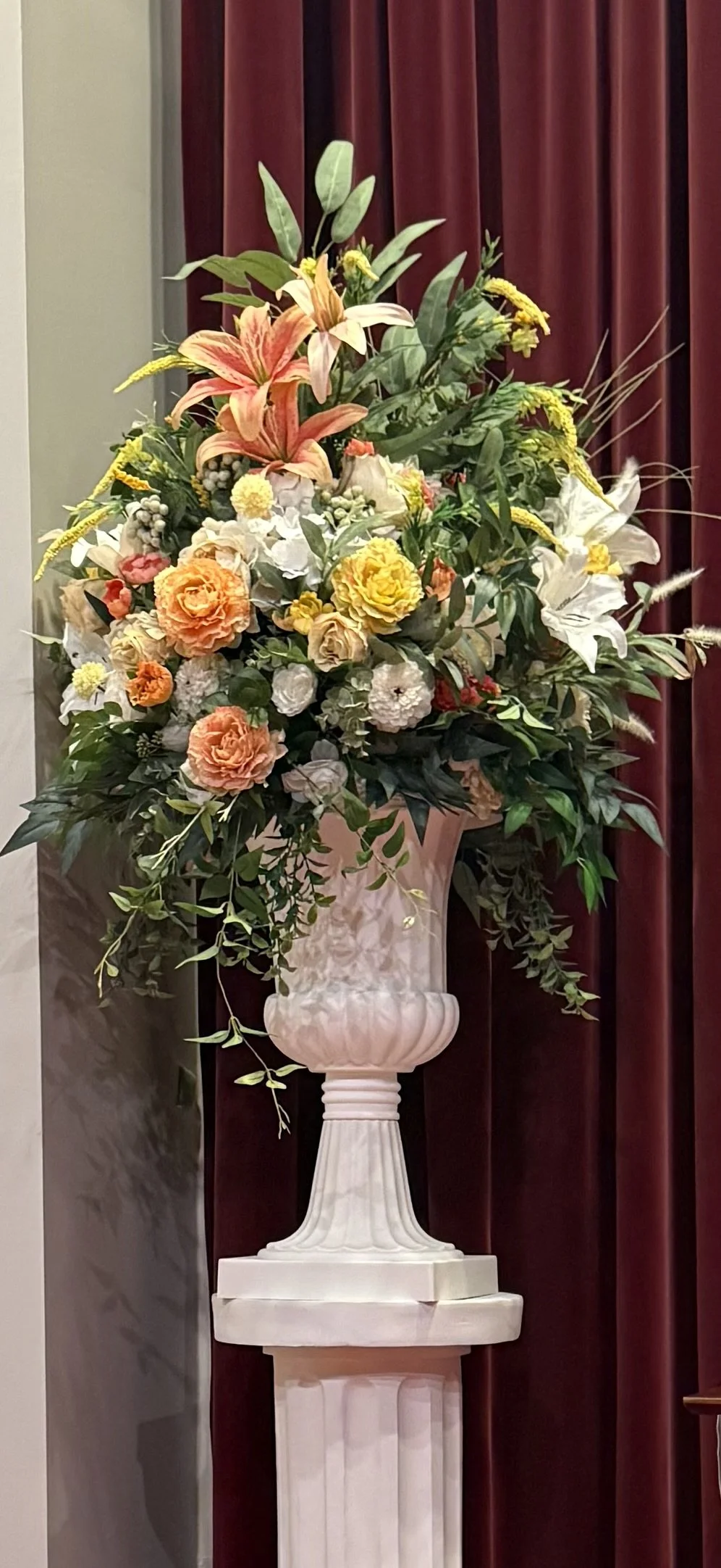 A large floral arrangement with pink lilies, orange and yellow roses, white lilies, and various greenery in a classical white urn.